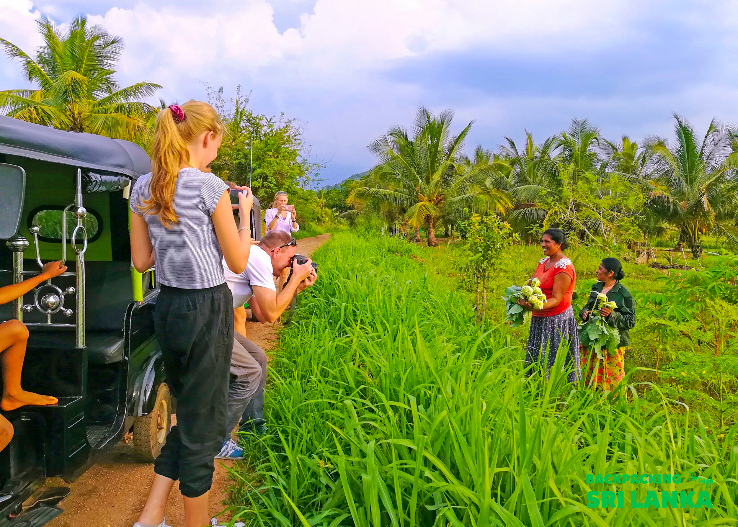 Backpackers photograph local women holding fresh produce next to a tuk-tuk on a rural path.