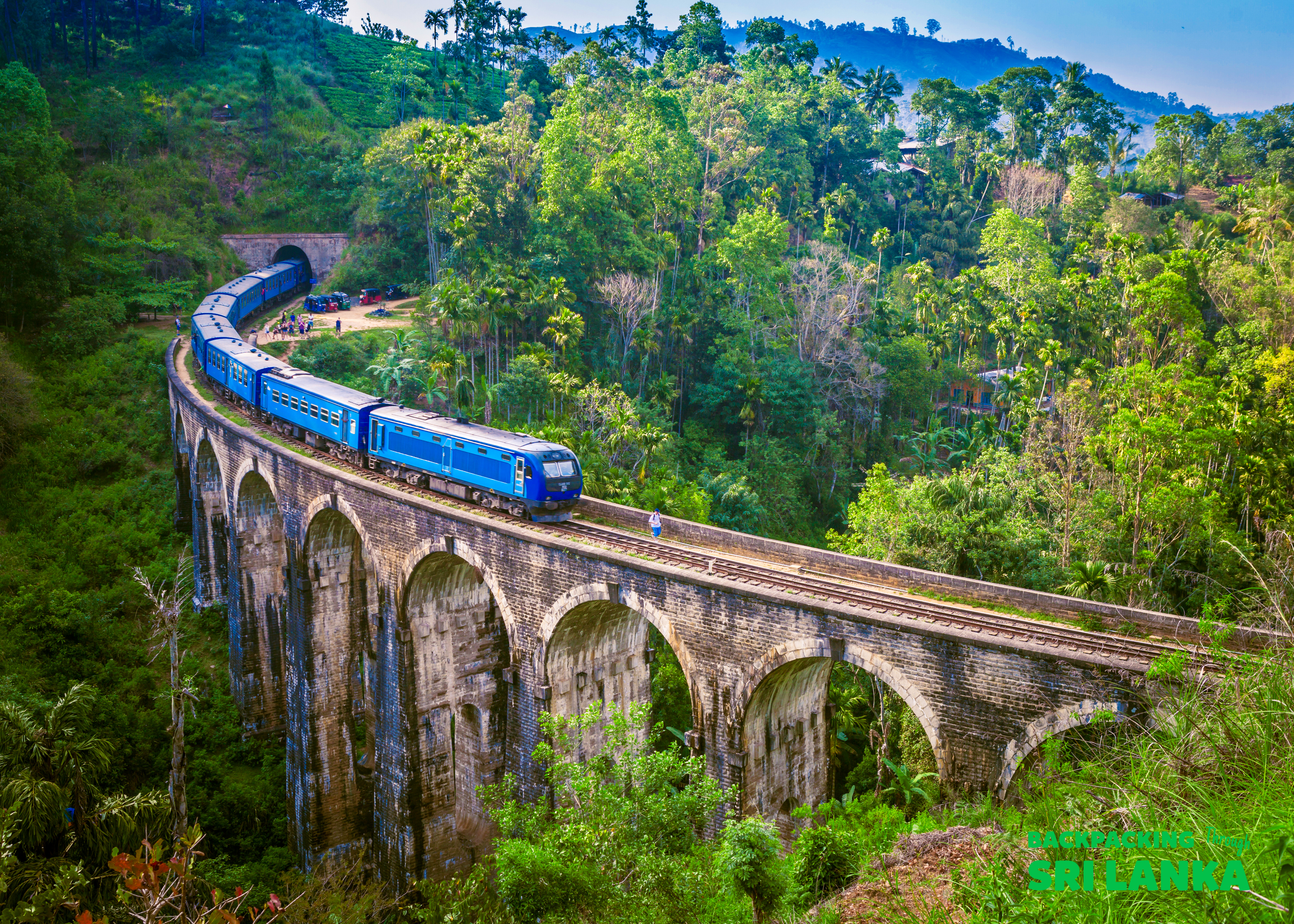 A blue train crosses the iconic Nine Arch Bridge surrounded by lush green mountains in Ella, Sri Lanka.