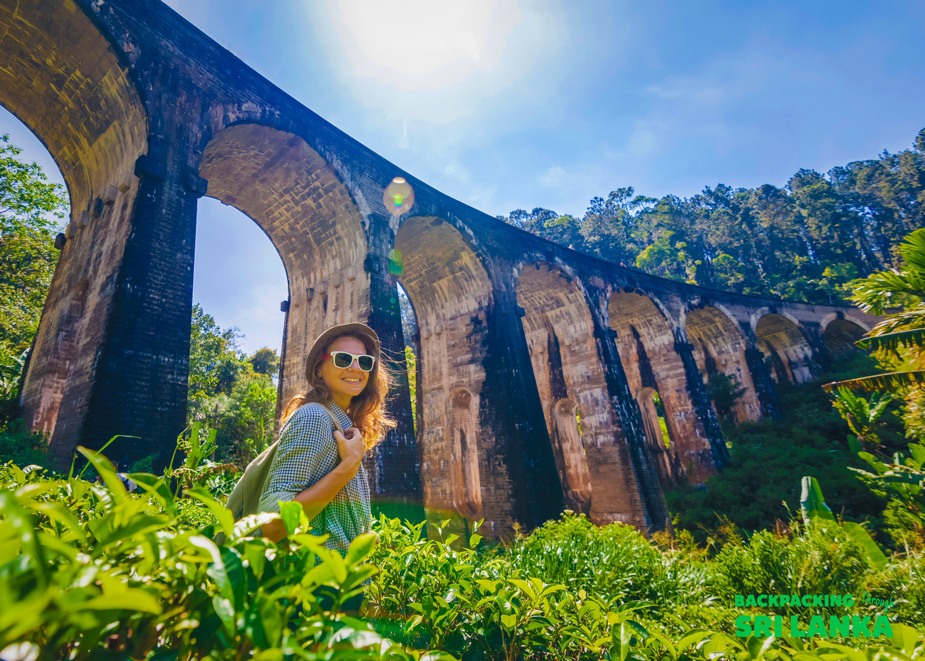 A smiling woman in sunglasses stands among lush tea plantations with the iconic 9 Arch Bridge in Ella.
