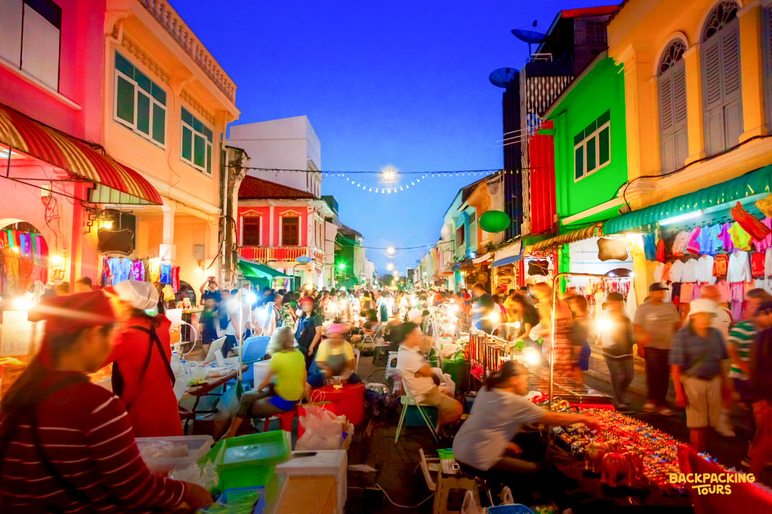 Shoppers explore a bustling night market in Phuket, featuring colorful buildings and bright stalls.