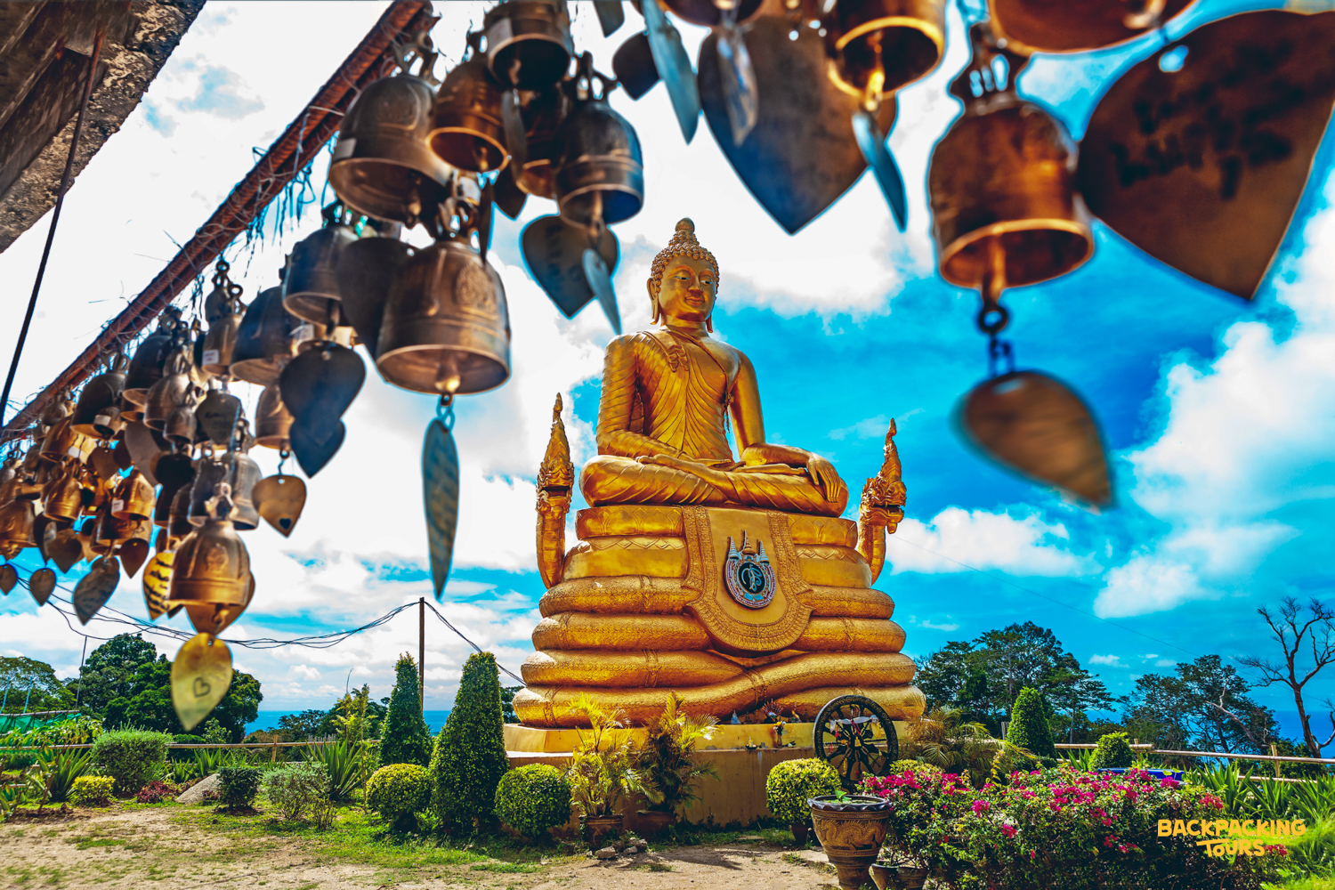 A majestic golden Buddha statue overlooks a scenic landscape, framed by hanging bells in Phuket.