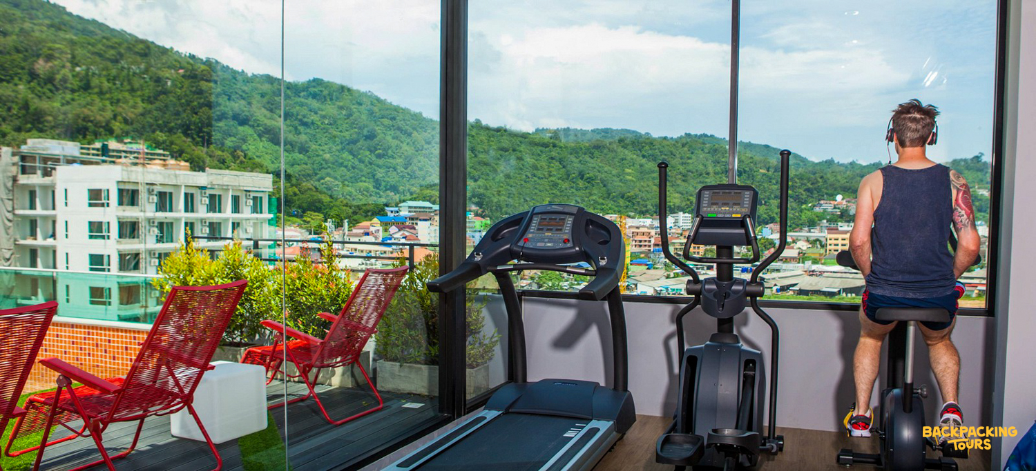A man exercises on a bike in a gym with a panoramic view of Phuket's lush hills.