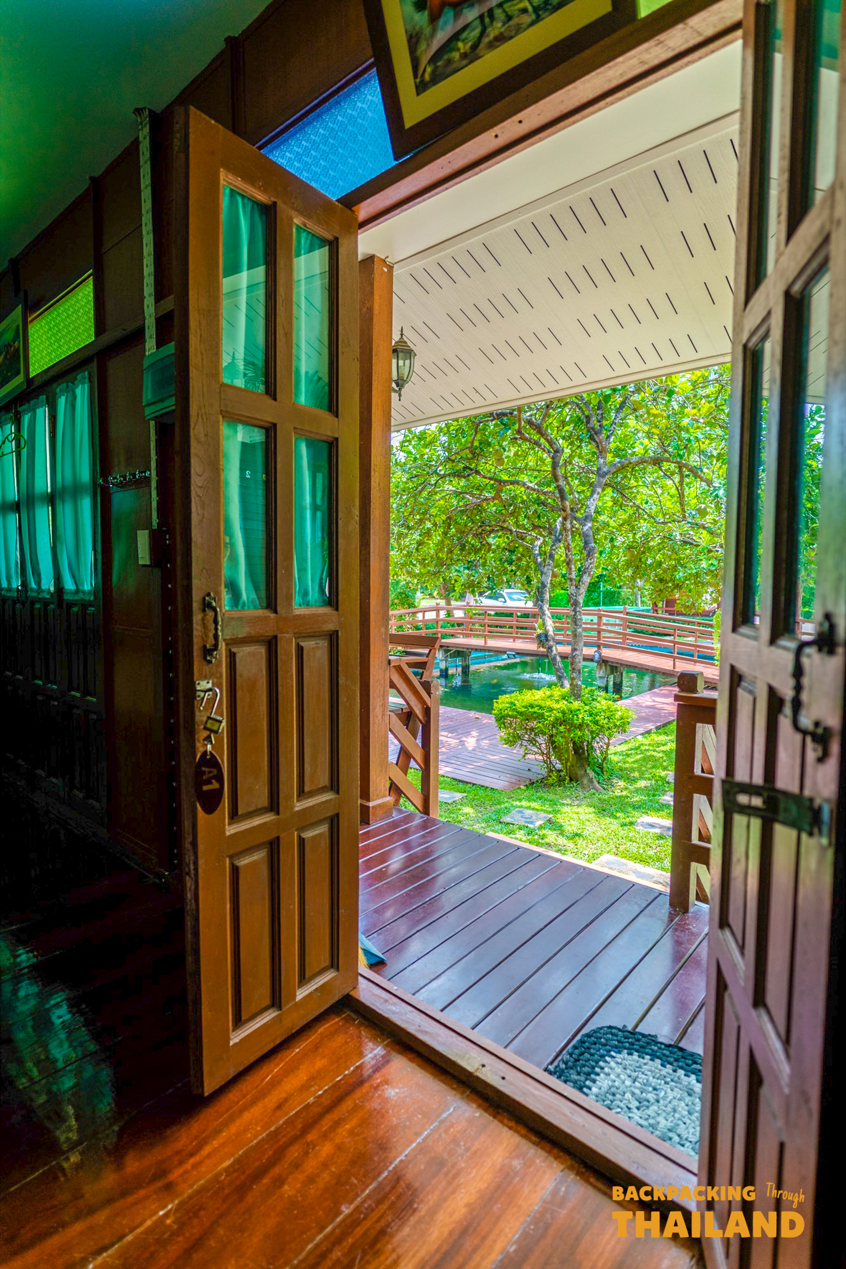Open wooden door reveals a tranquil homestay view with a bridge over water in Amphawa.