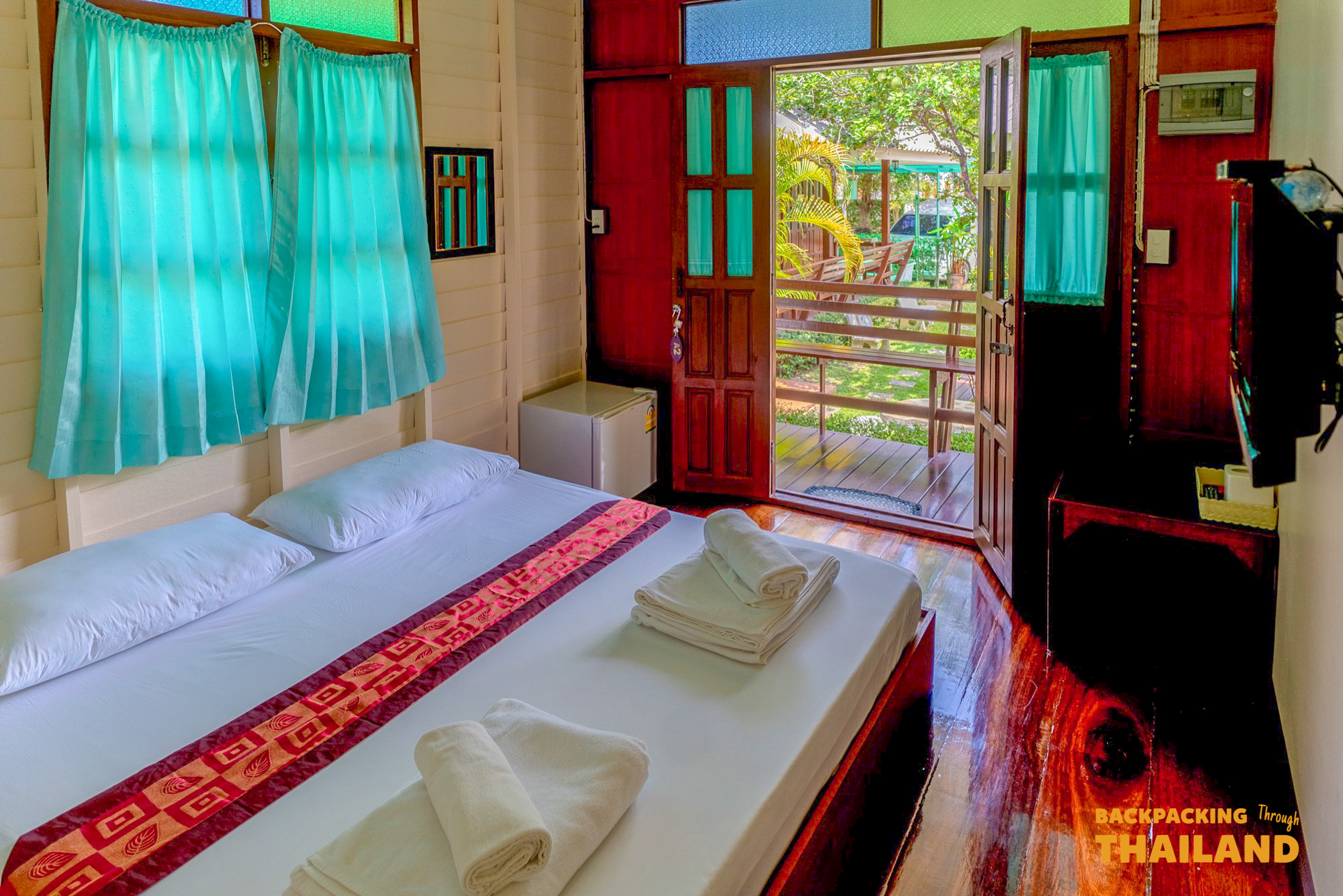 A comfortable homestay bedroom in Amphawa, Thailand, with a bed, fresh towels, and a green outdoor view.