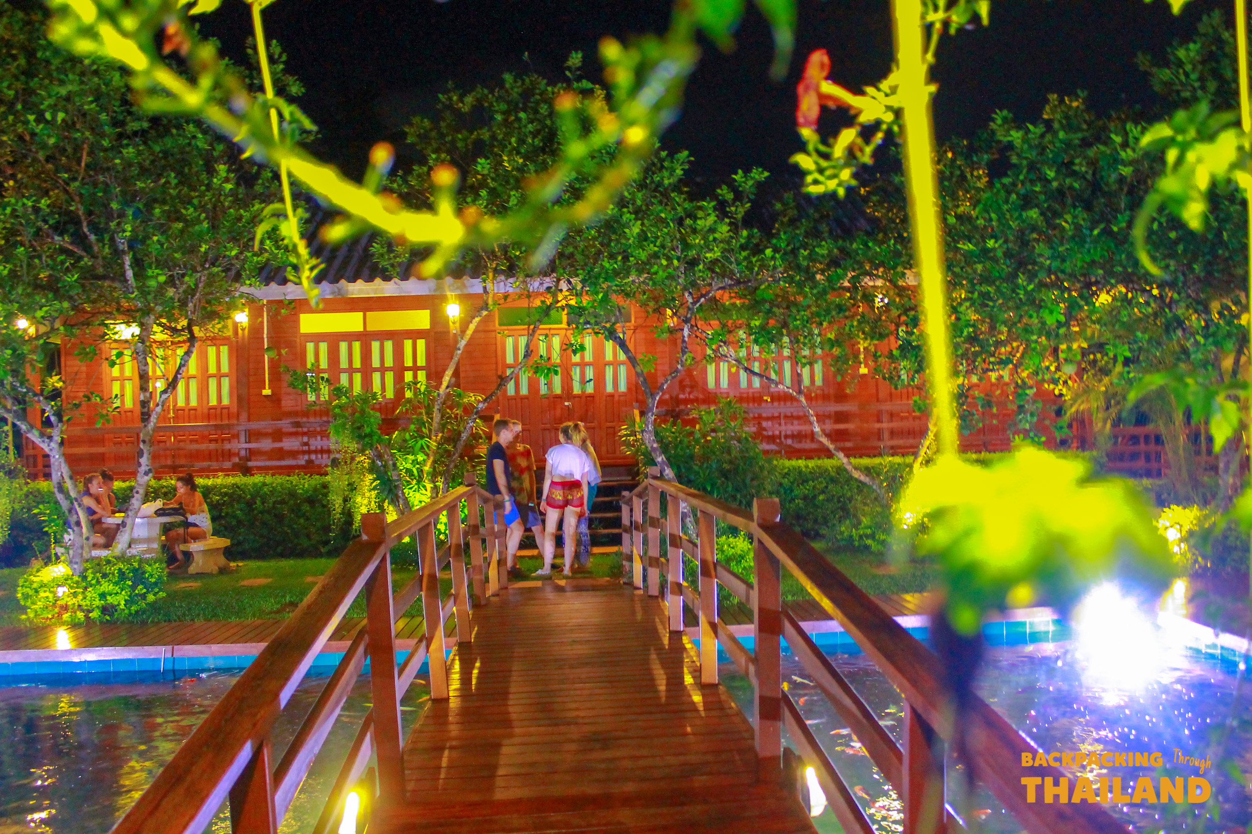 Tourists cross a wooden bridge towards a charming homestay lit up at night in Amphawa, Thailand.