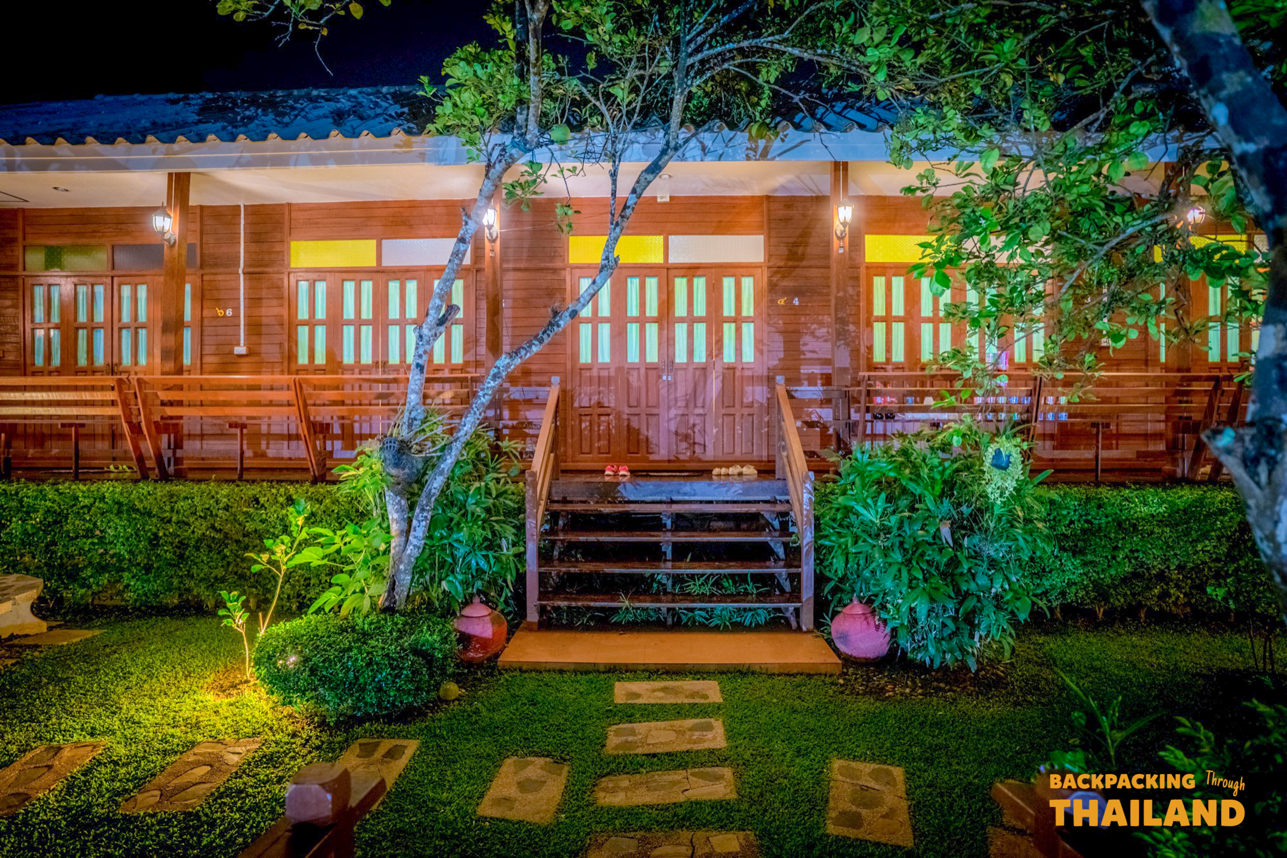 A charming traditional wooden homestay in Amphawa, Thailand, glowing warmly at night with a lush garden.