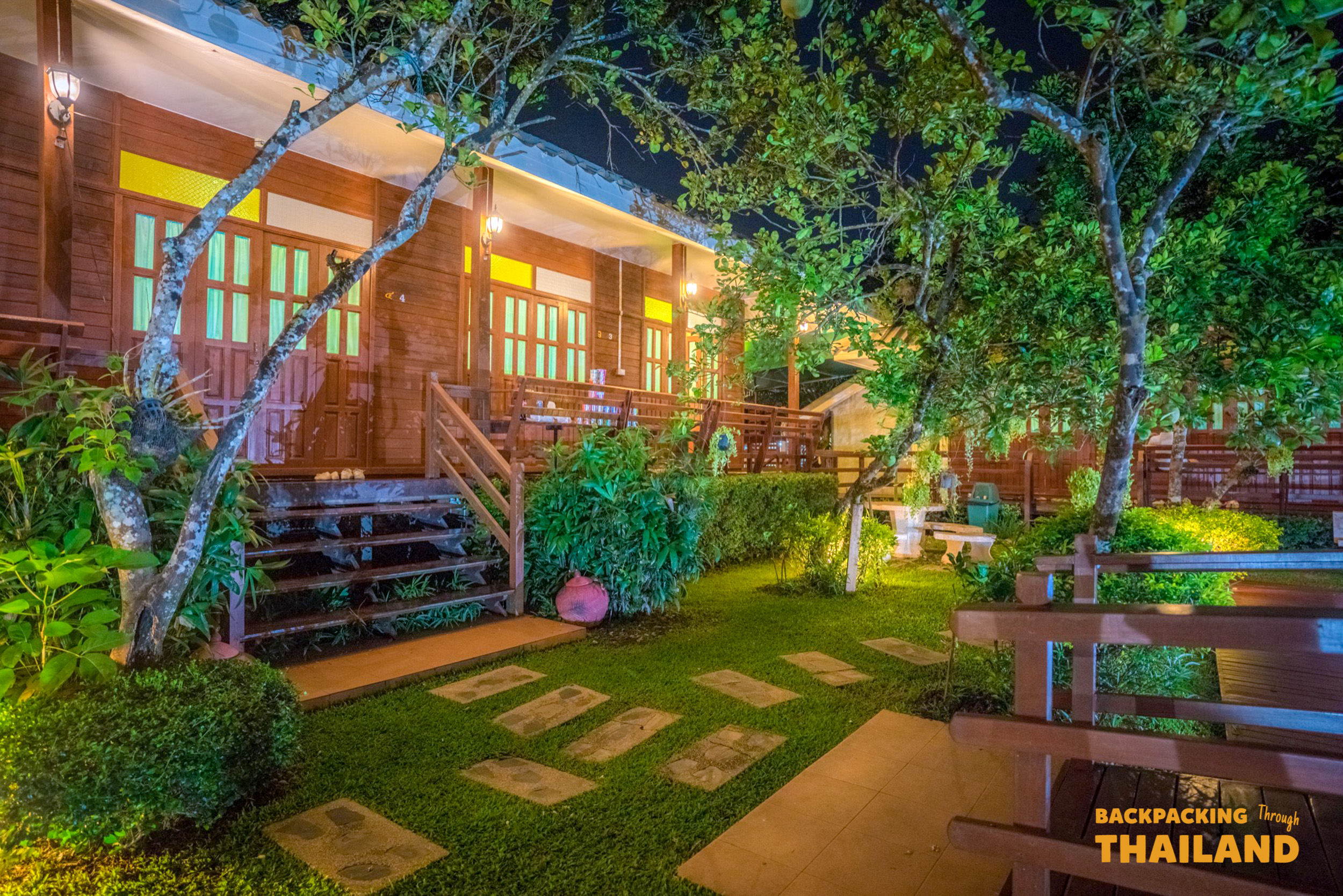 Cozy wooden homestay in Amphawa, Thailand, illuminated at night with a vibrant green garden.