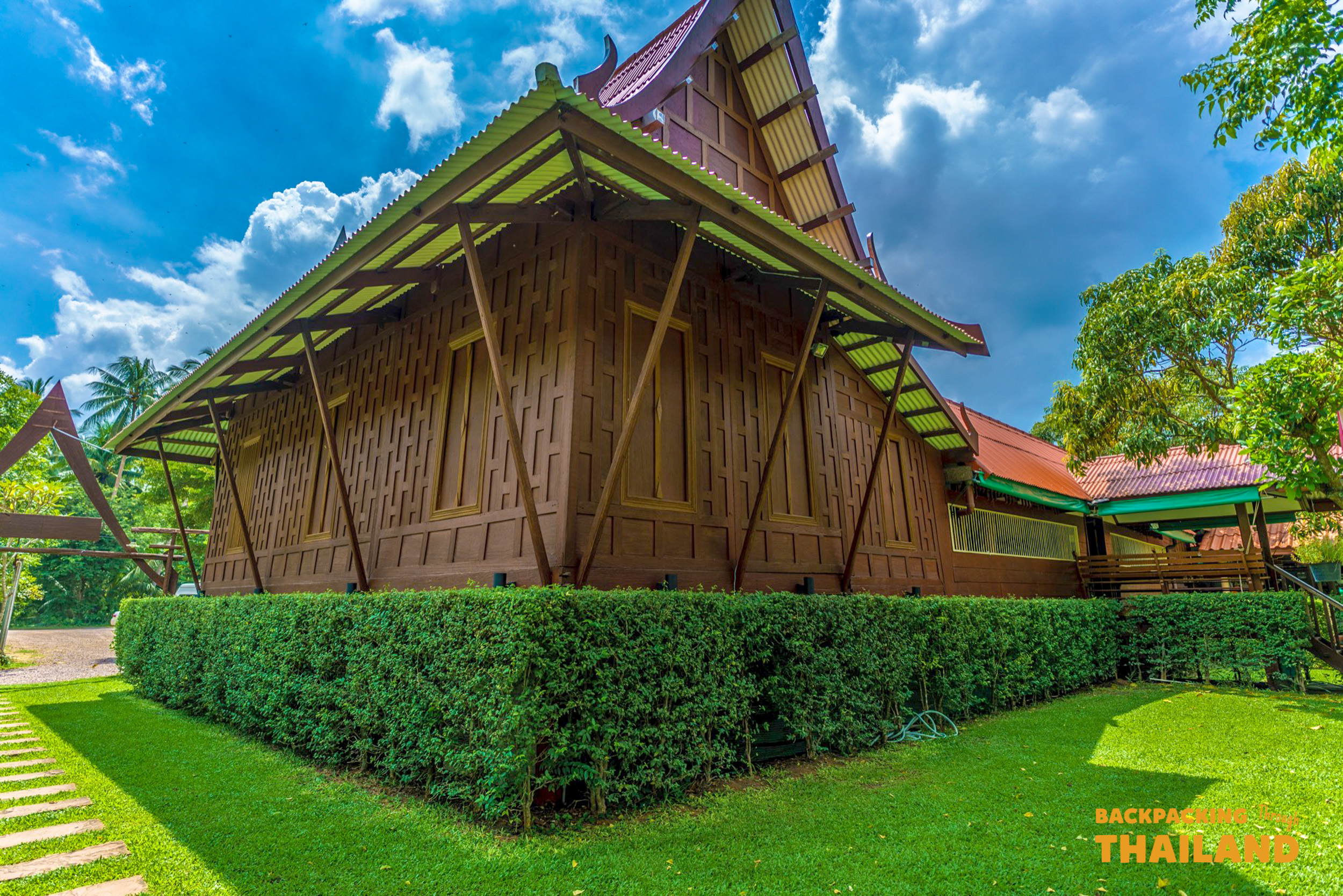 A beautiful traditional wooden homestay building with green hedges and a lush lawn in Amphawa.