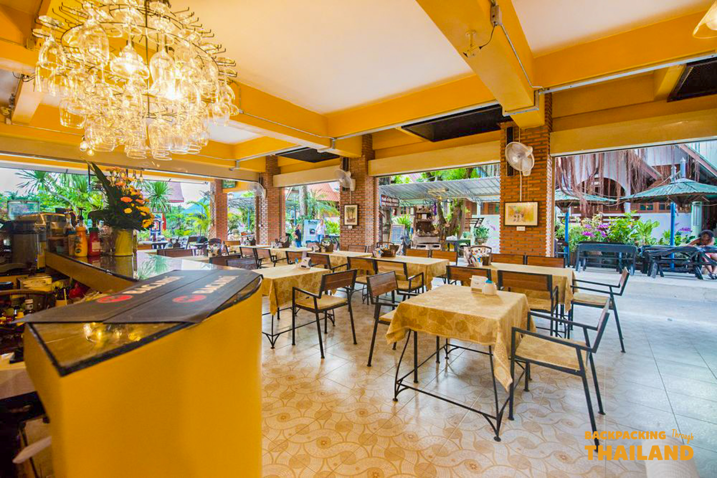 A bright and inviting open-air restaurant in Kanchanaburi, Thailand, with yellow walls and a sparkling chandelier.