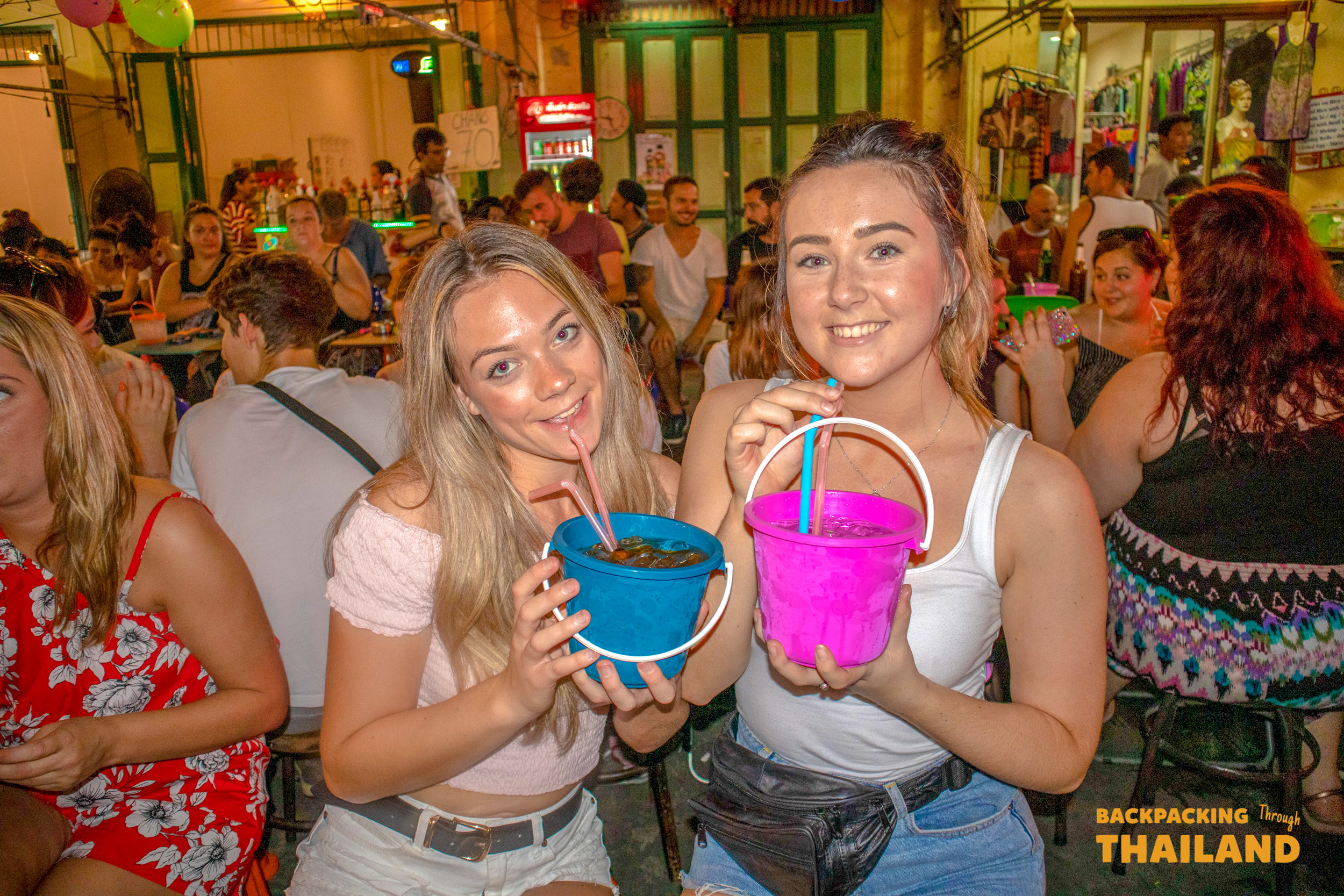 Two happy women enjoying colorful bucket drinks on a lively street in Bangkok at night.