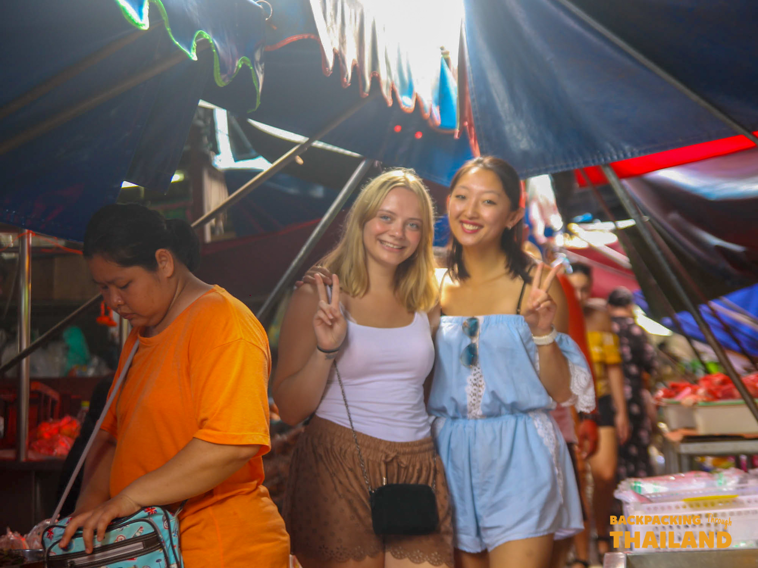 Two happy women pose with peace signs at a bustling market in Amphawa, Thailand.