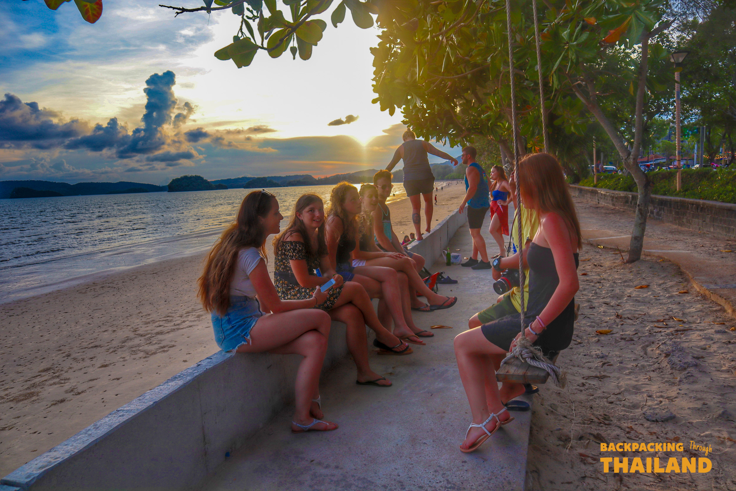 Young travelers relax on a beach wall, enjoying a beautiful sunset over the ocean in Krabi, Thailand.