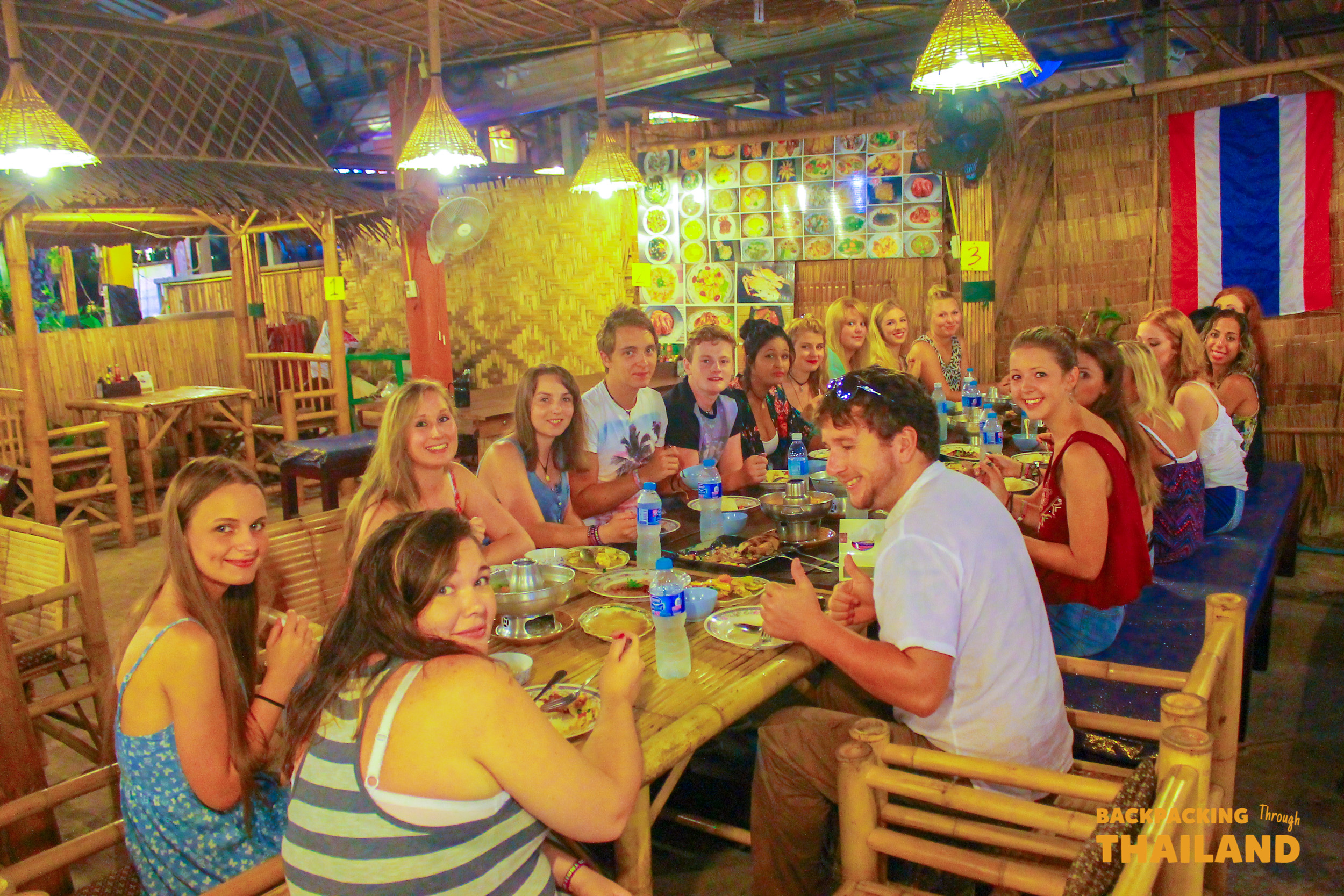 A large group of young backpackers enjoying a lively dinner together at a rustic restaurant in Krabi, Thailand.