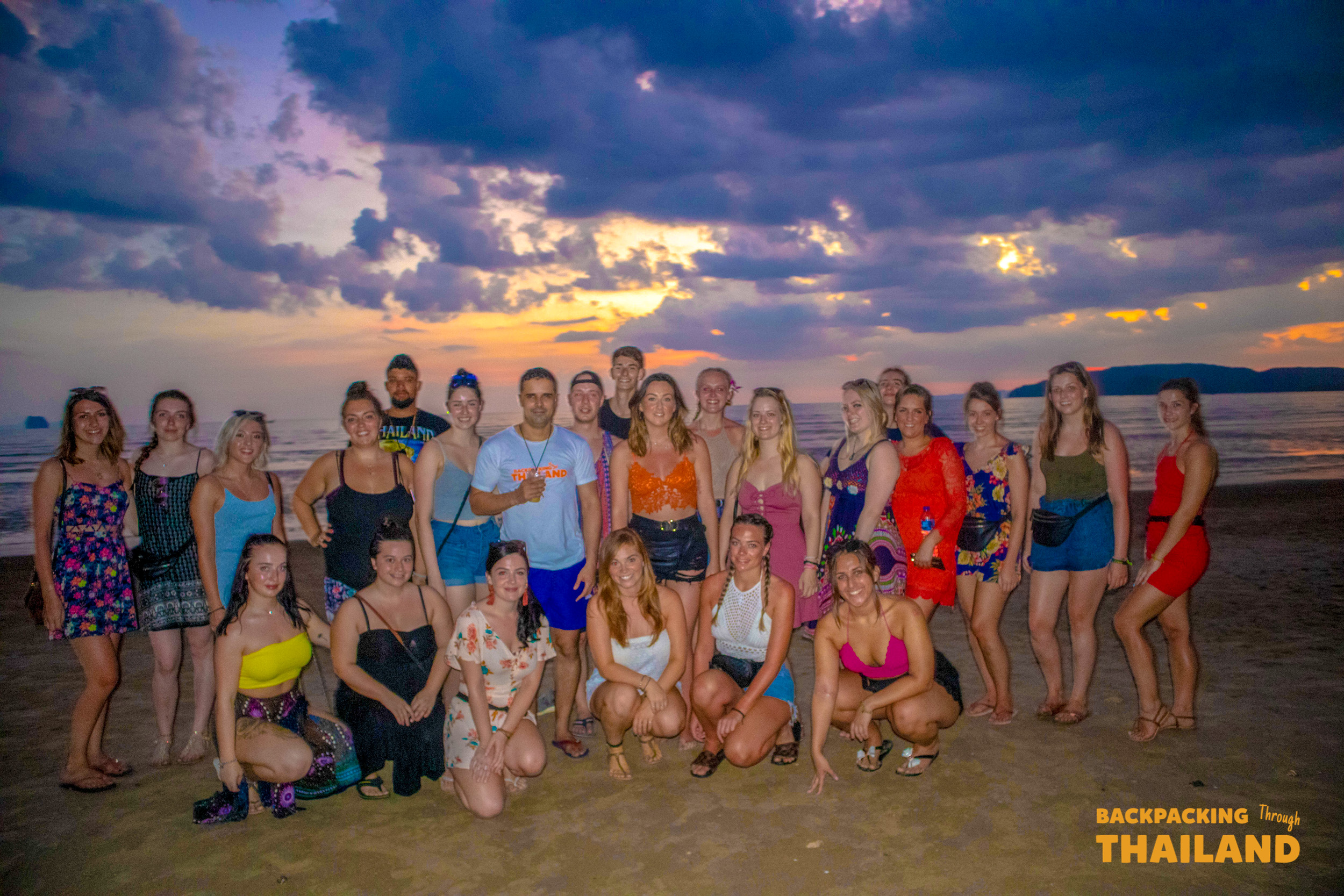 A large group of happy backpackers smiles for a photo on a beautiful Krabi beach at sunset.
