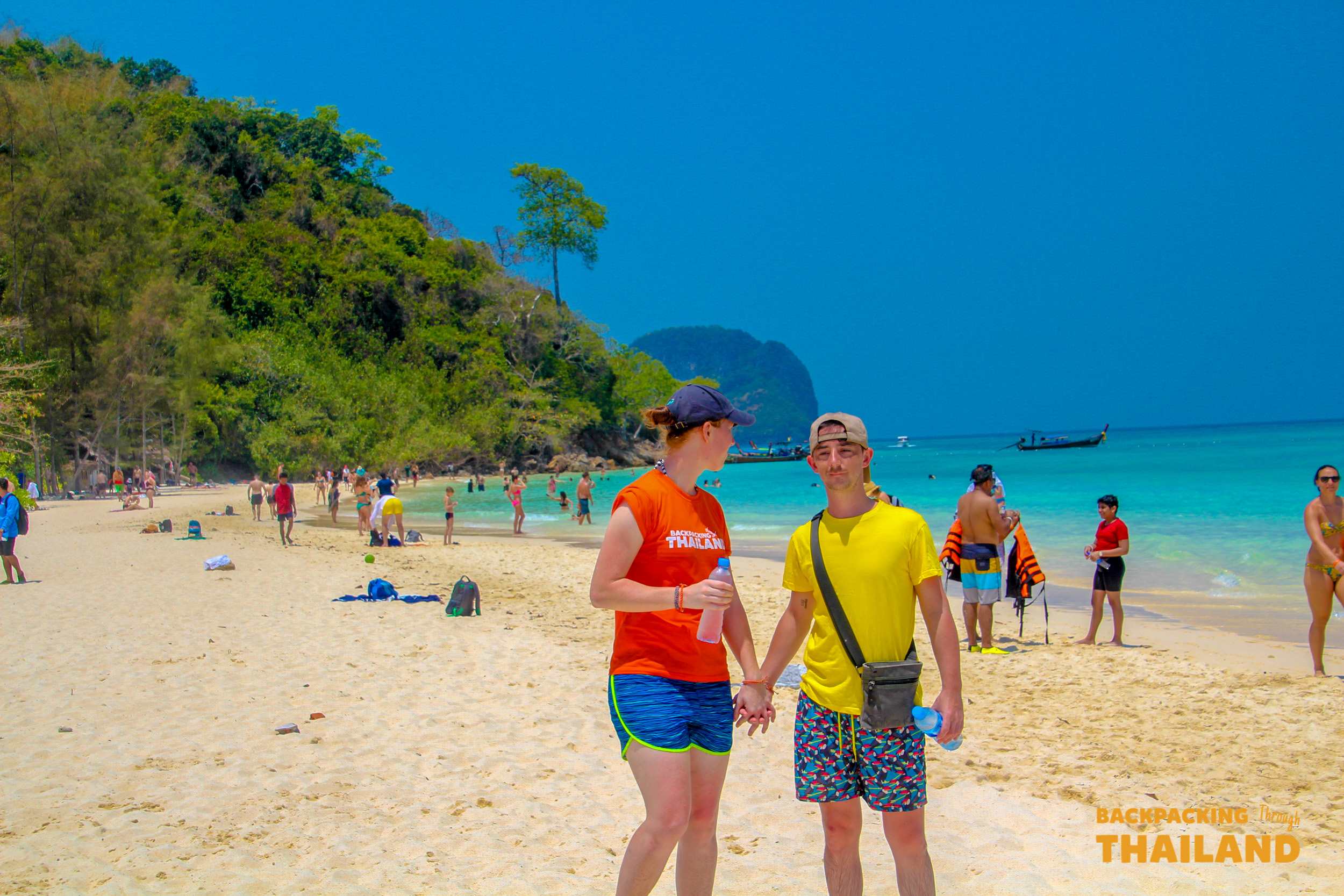 A young couple holding hands on a sunny Krabi beach in Thailand, with turquoise water.