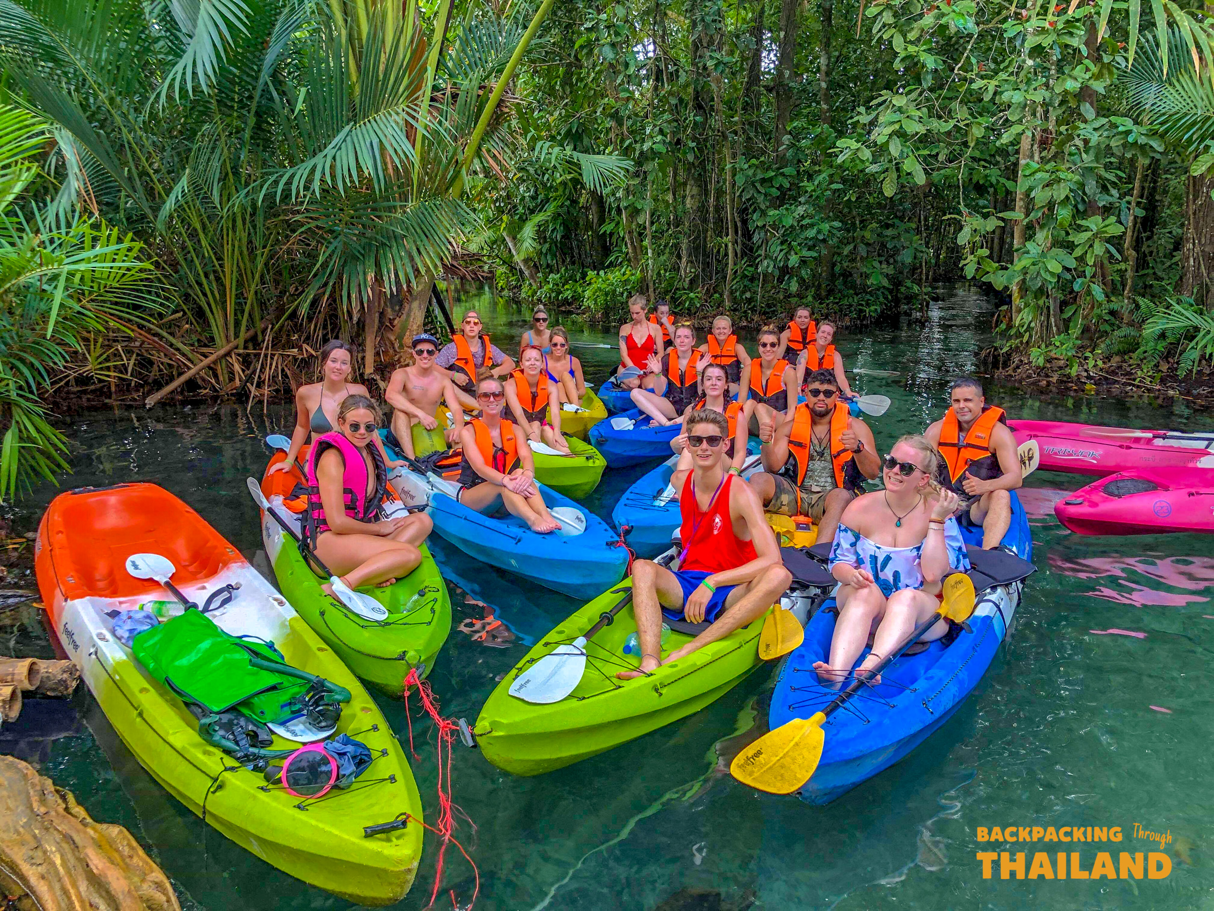 A group of happy backpackers kayaking on clear emerald water through a lush jungle in Krabi, Thailand.
