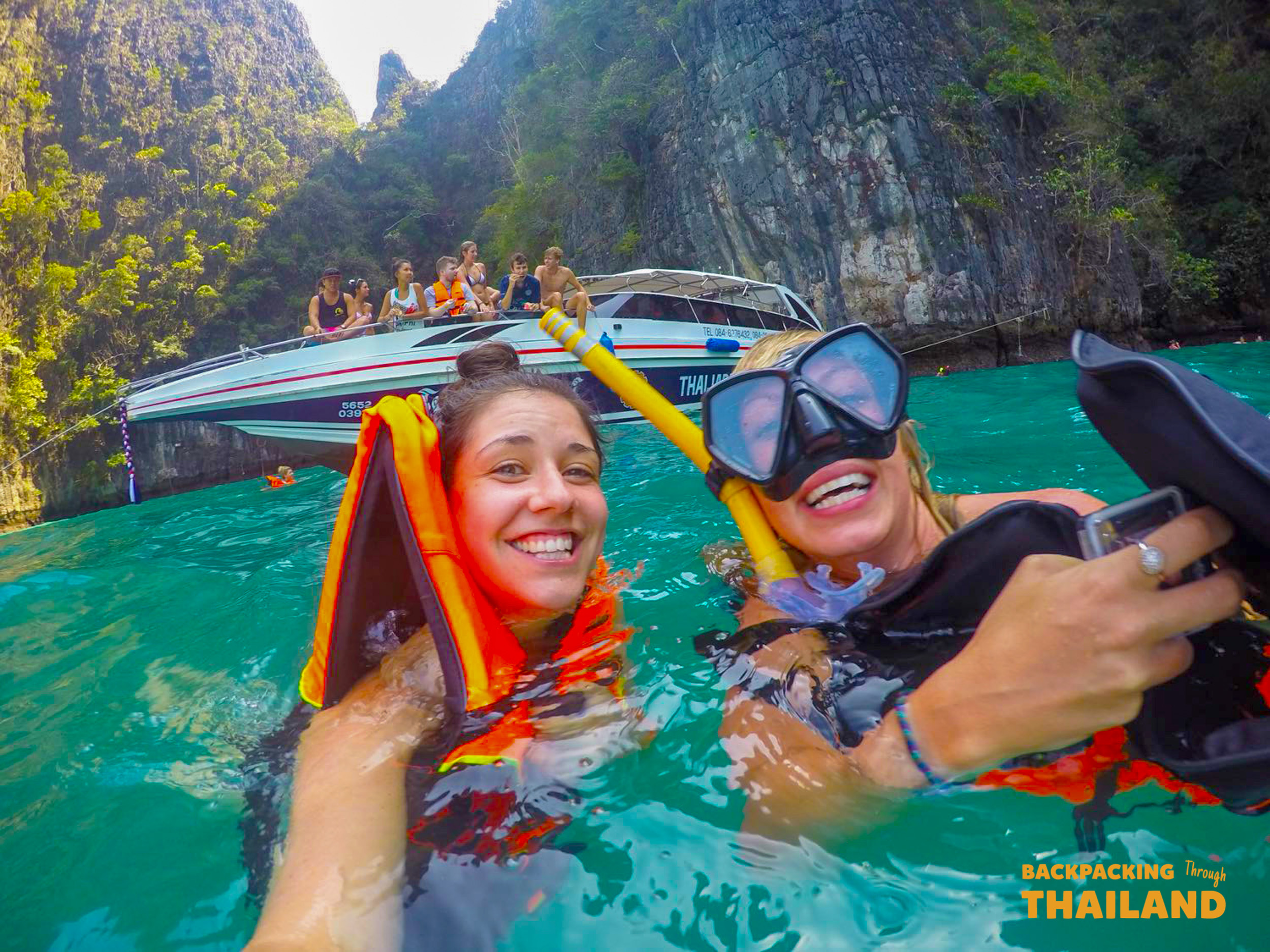 Two happy women snorkel in the clear turquoise water near towering limestone cliffs in Thailand.