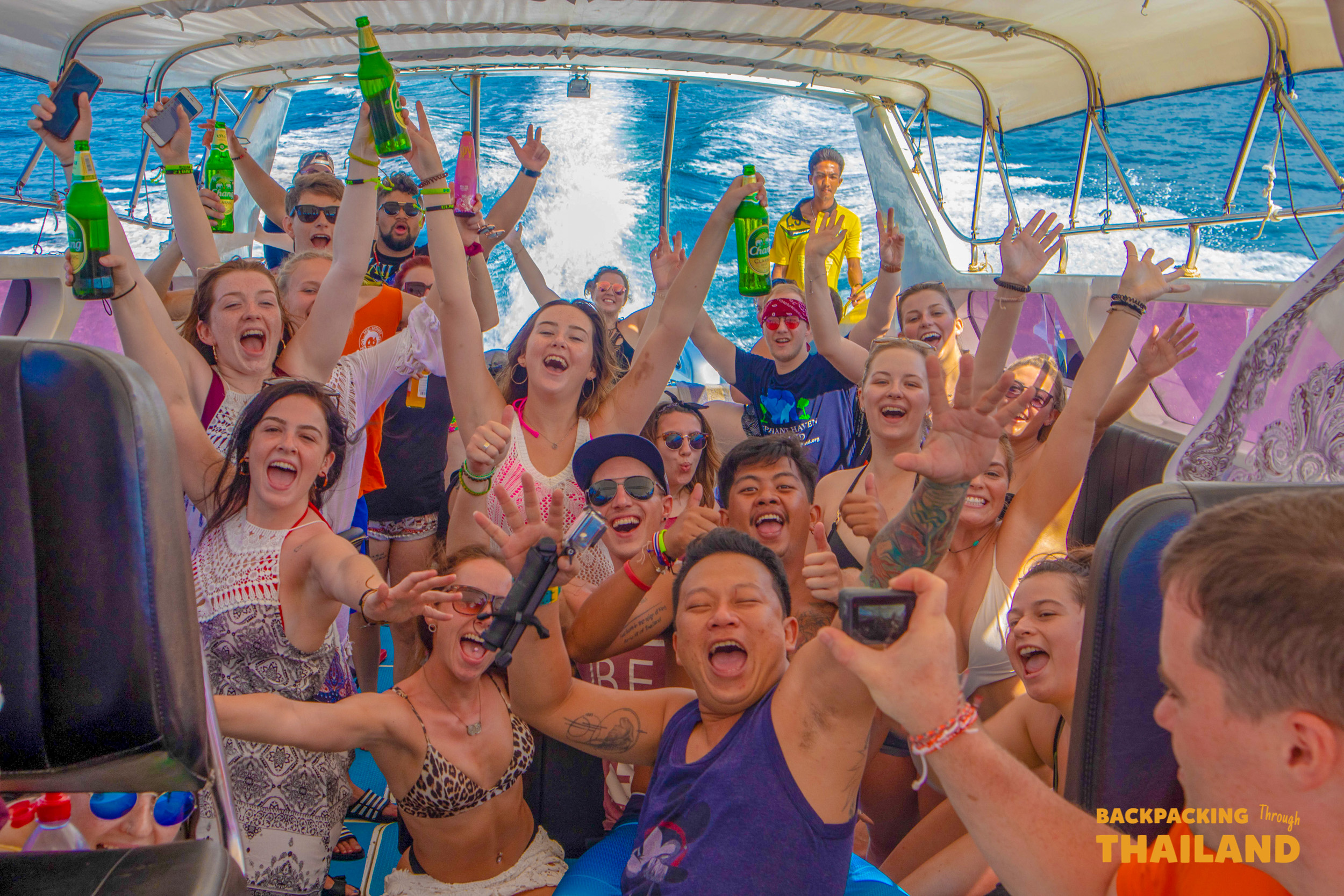 Joyful backpackers cheer and raise bottles on a speed boat in the beautiful blue waters of Thailand.