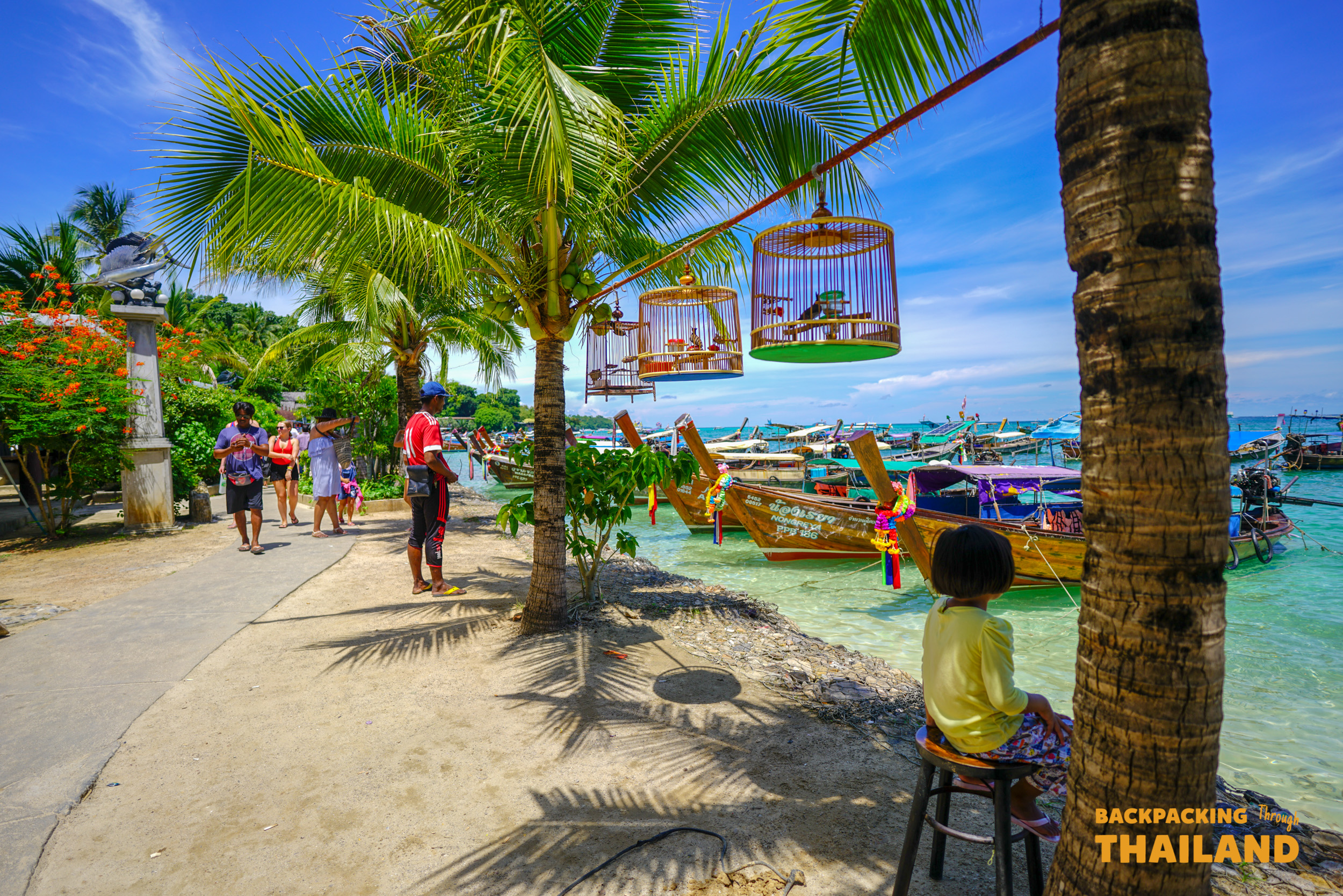 Tourists walk along a sunny Koh Phi Phi beach with hanging birdcages and traditional longtail boats.