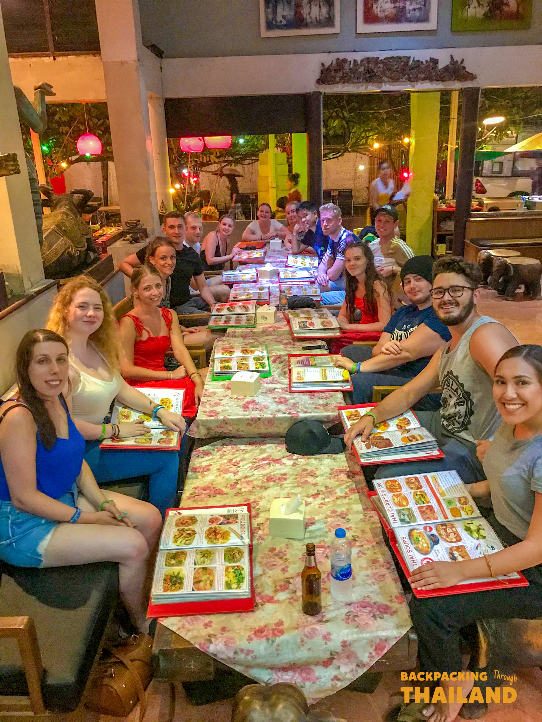 A large group of happy backpackers enjoys a lively dinner together at an outdoor restaurant in Thailand.