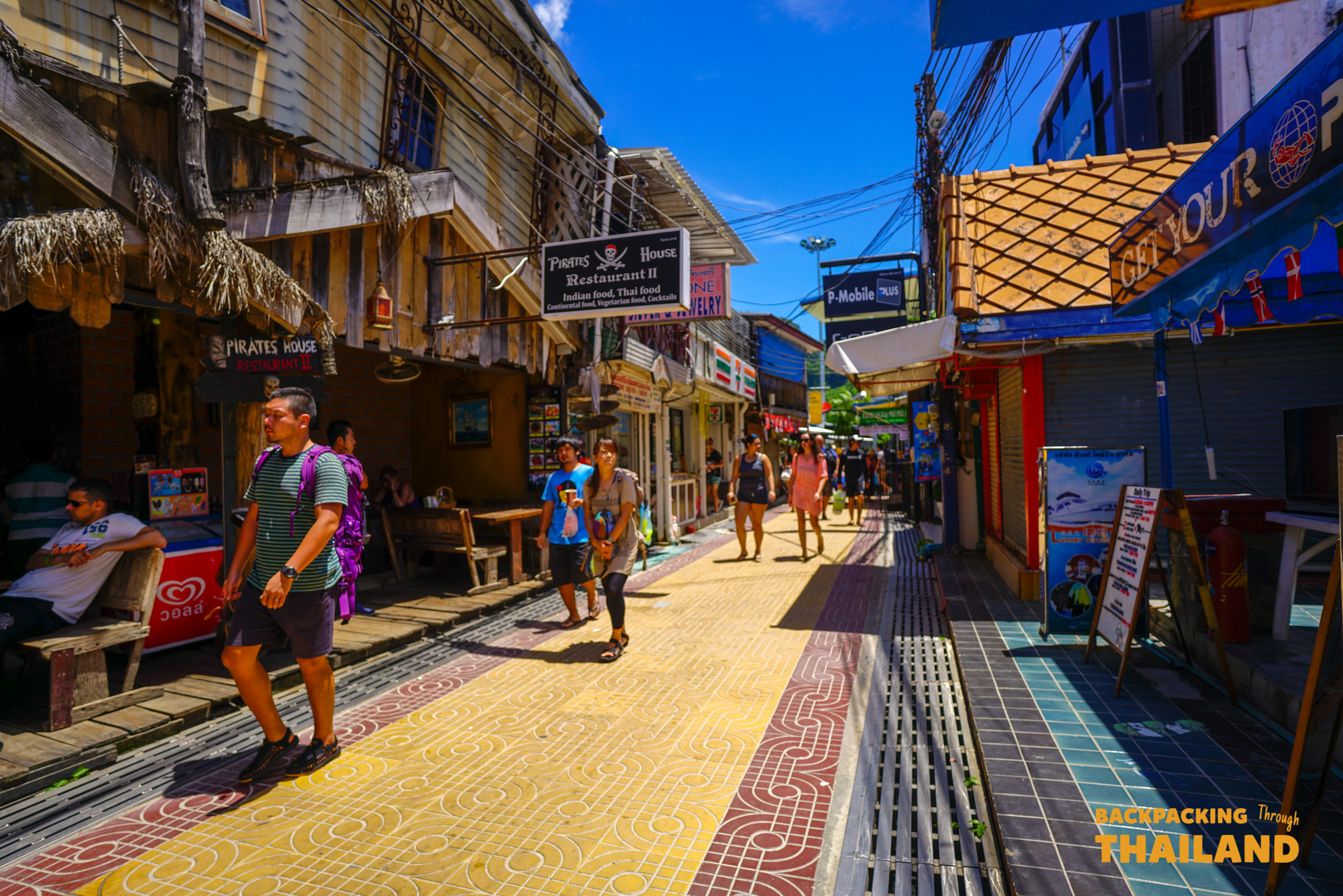 Travelers explore the vibrant, narrow streets of Koh Phi Phi, browsing shops and enjoying the sunny day.