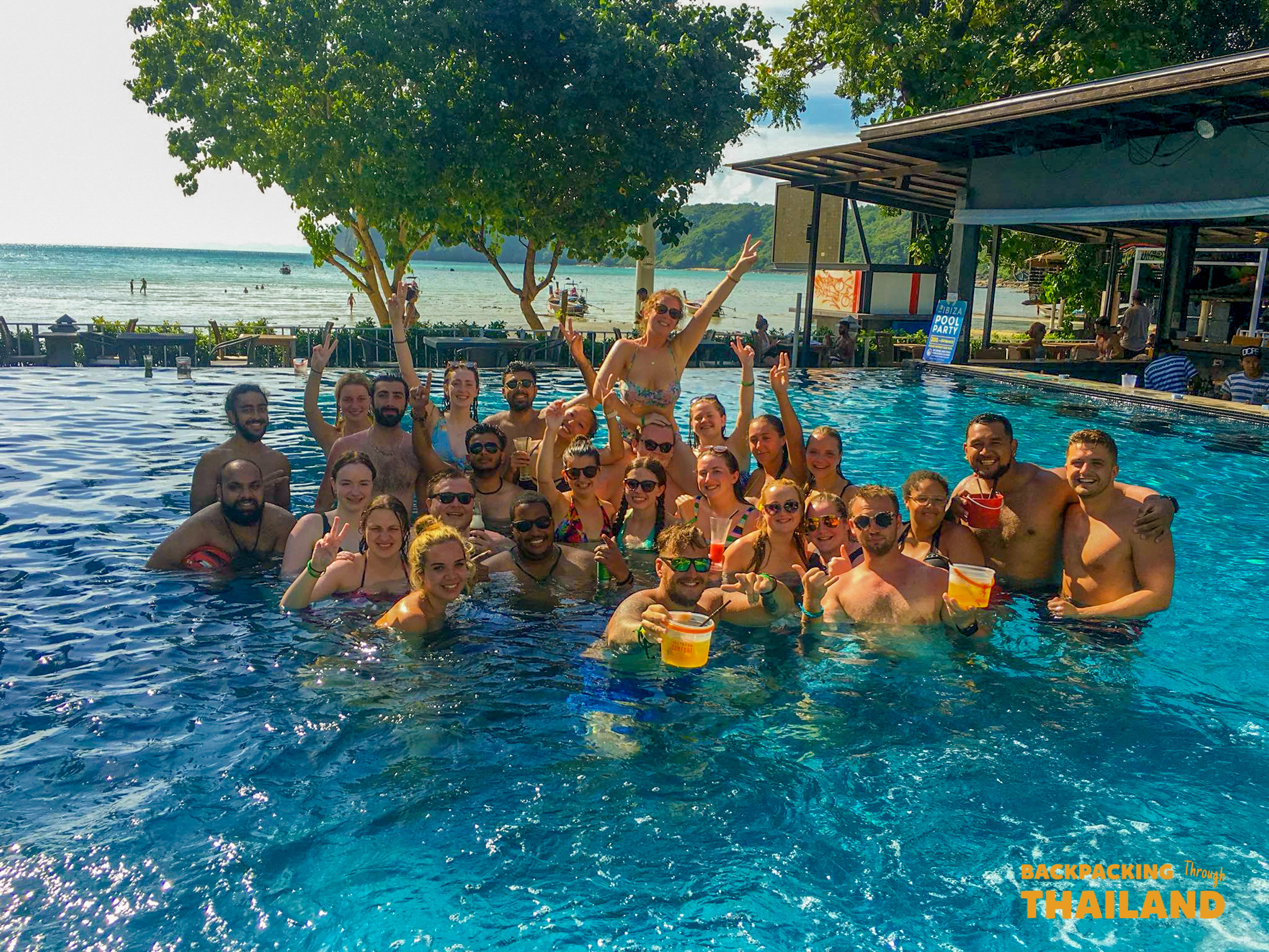 Smiling backpackers celebrating in a lively infinity pool with tropical ocean views in Thailand.
