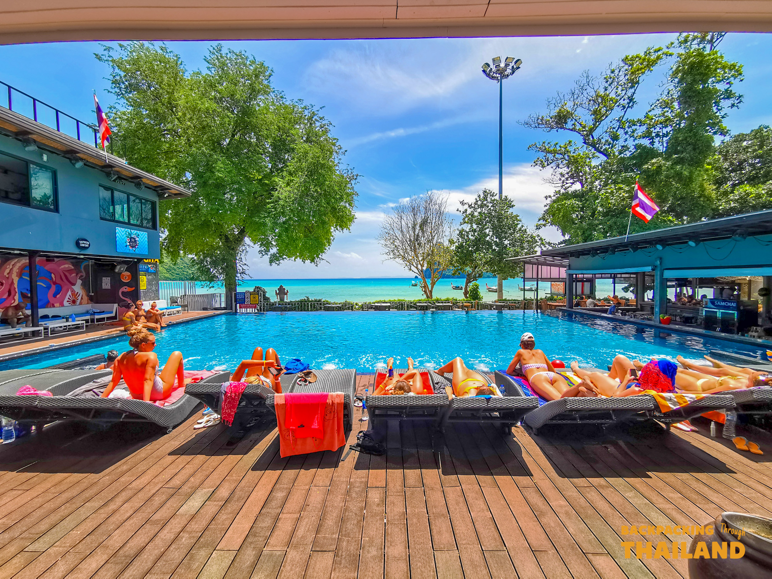 Backpackers relax by a vibrant blue pool overlooking the clear turquoise ocean on Koh Phi Phi island.