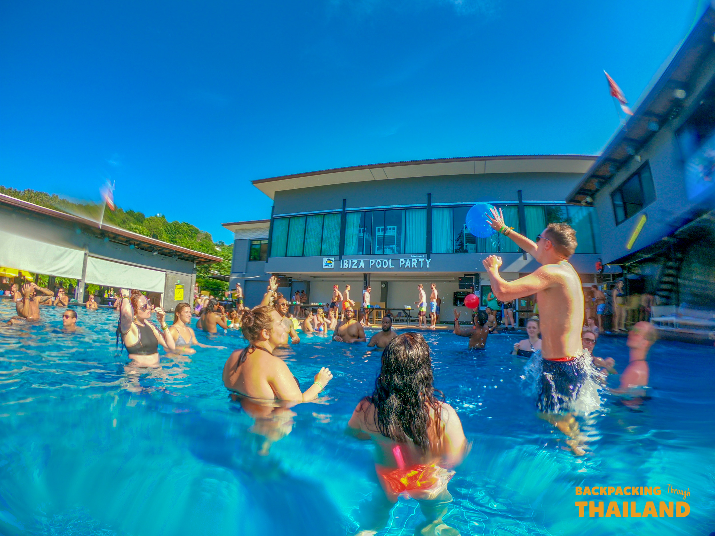 Young travelers have fun at a vibrant Ibiza-themed pool party under a sunny sky.
