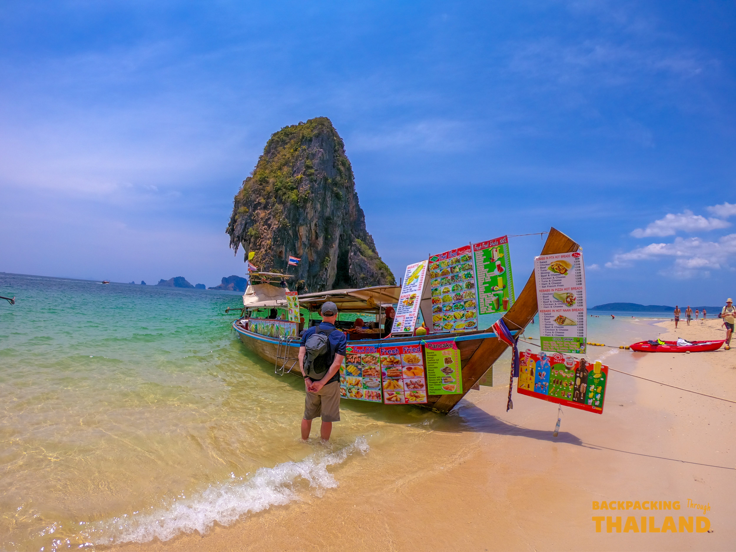 A traveler wades in clear water by a colorful longtail food boat at Railay Beach, Thailand.