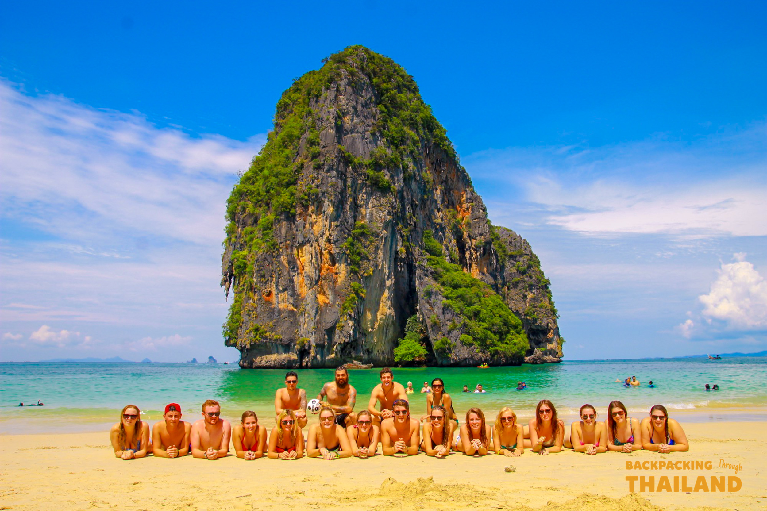 Smiling backpackers relax on Railay Beach's white sand with a towering limestone island in Thailand.
