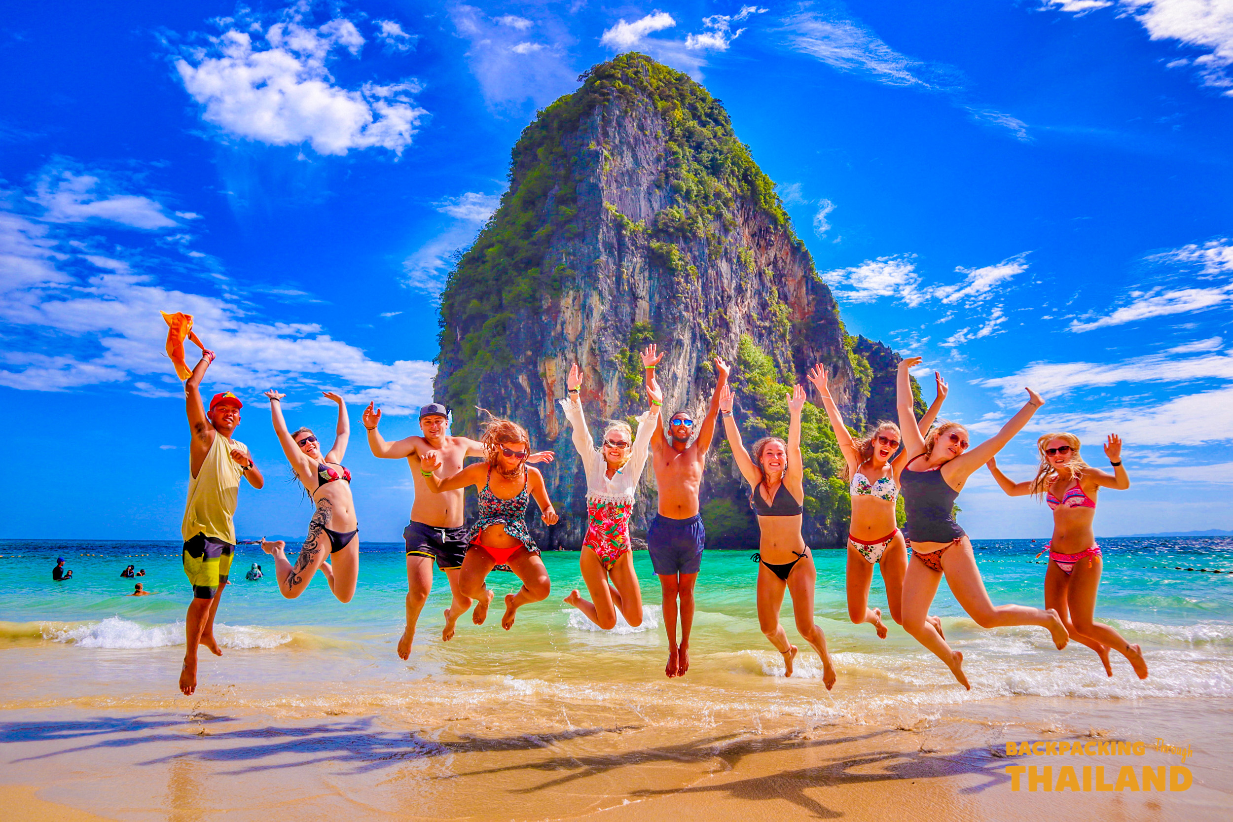 Joyful backpackers leap on the white sands of Railay Beach, Thailand, surrounded by limestone cliffs.