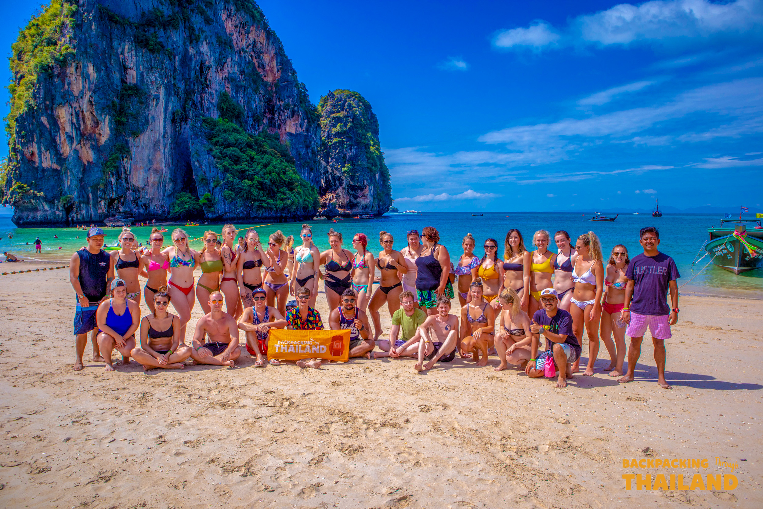 A large group of happy backpackers pose on the white sands of Railay Beach, Thailand.