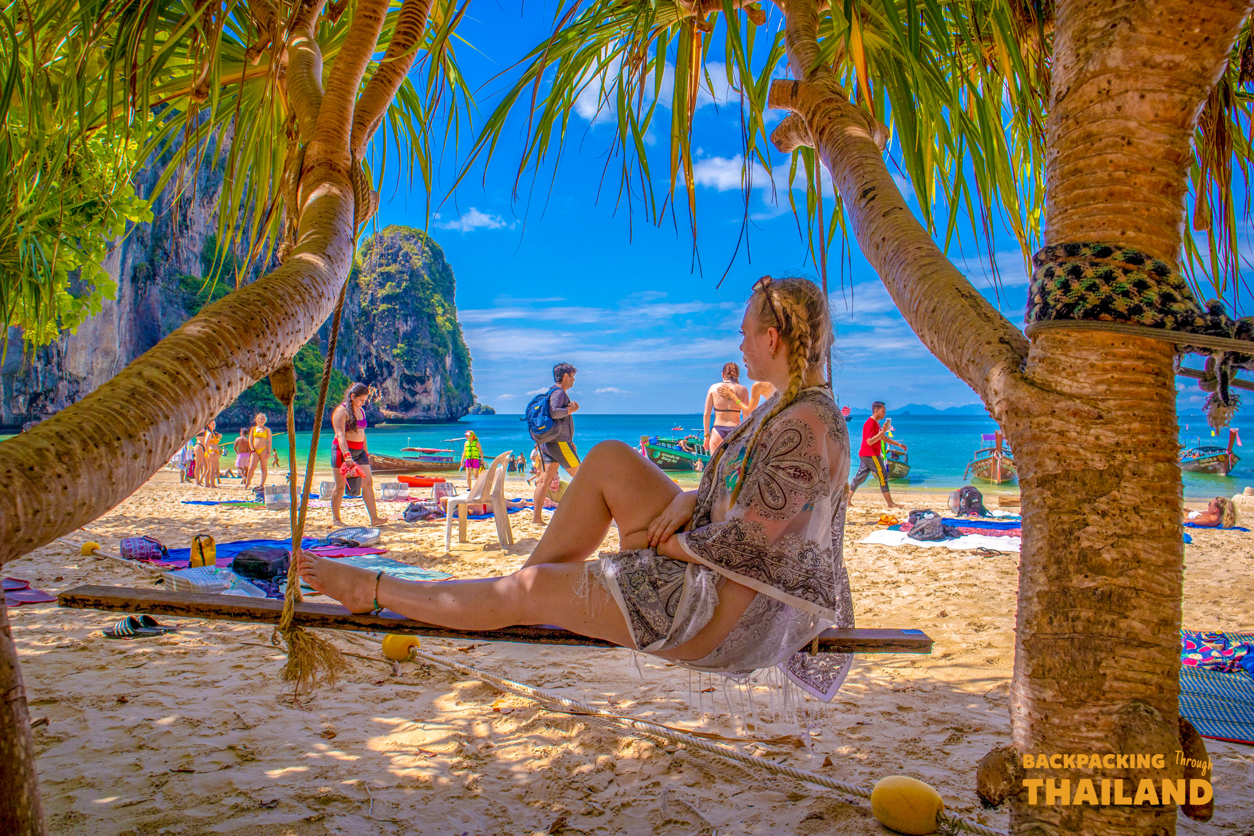 A woman enjoys a swing on the white sands of Railay Beach, Thailand, framed by palm trees.