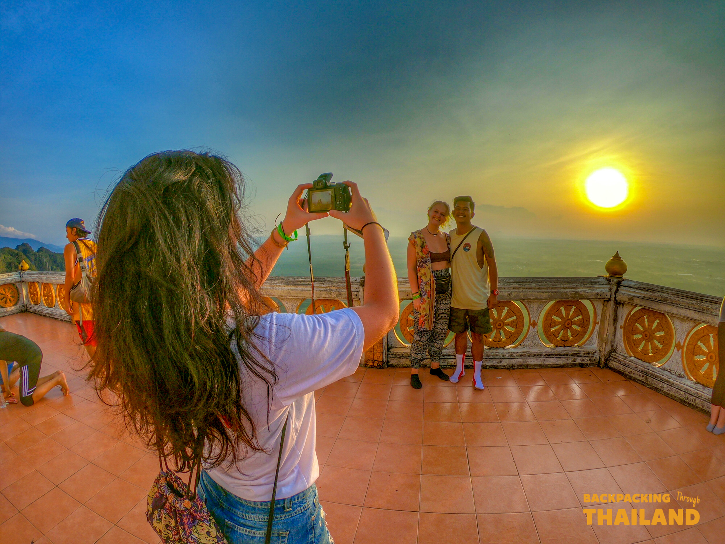 A woman photographs two friends posing on an ornate temple balcony at sunset in Krabi.