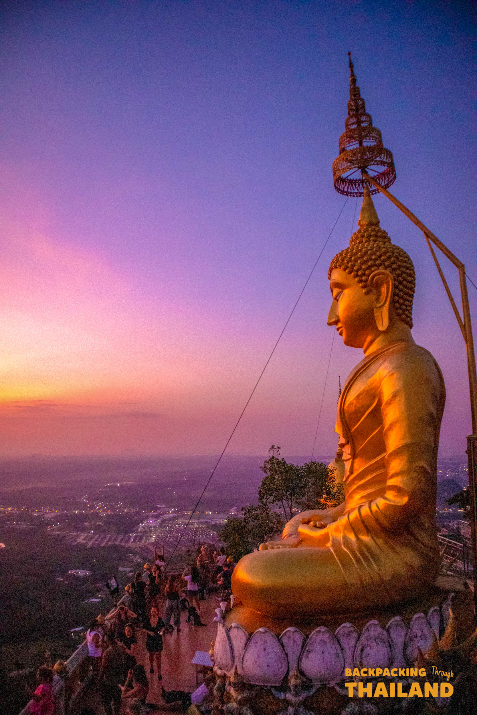 Golden Buddha statue at Tiger Cave Temple in Krabi, with people enjoying the stunning sunset view.