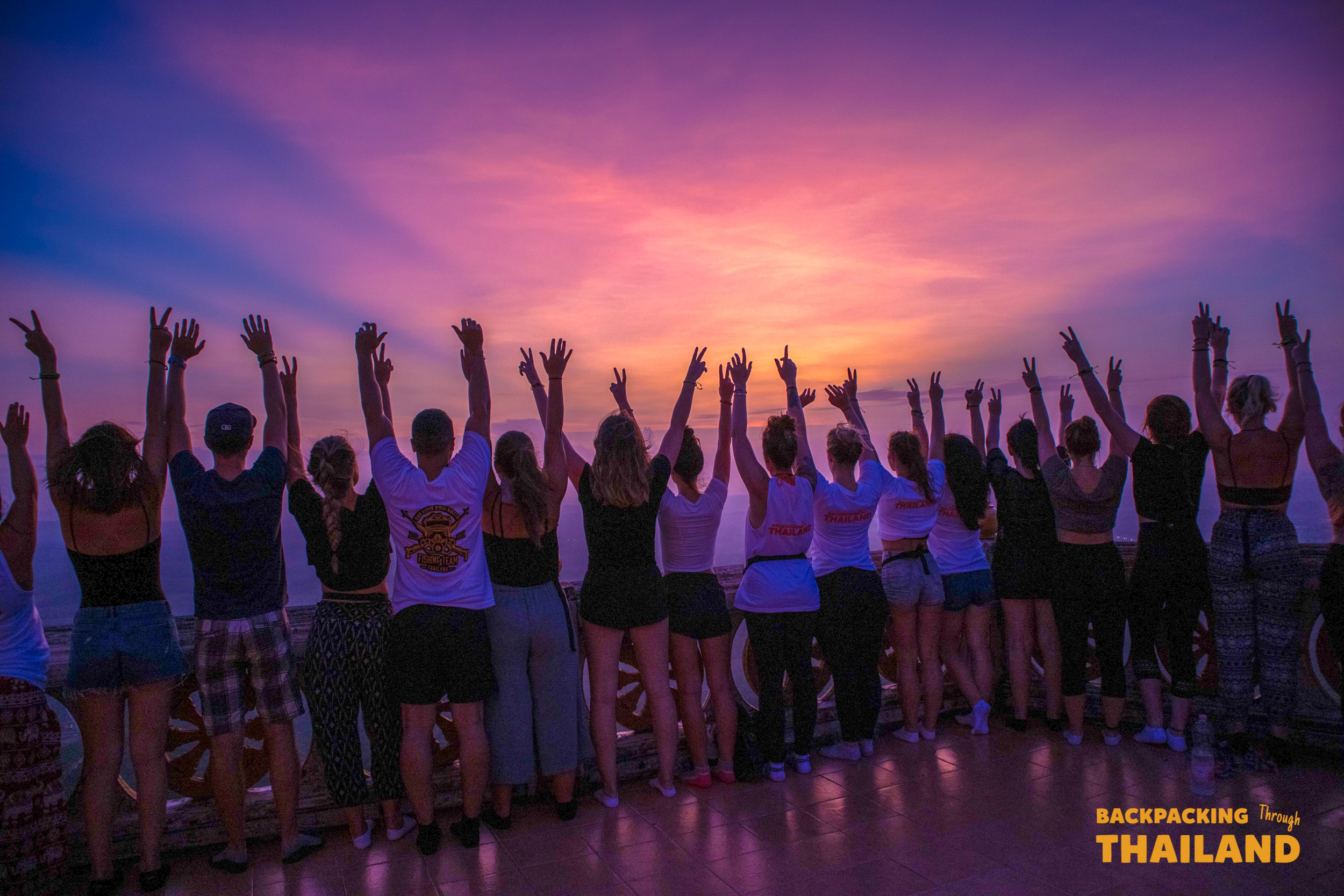 Backpackers raise their arms, celebrating a vibrant sunset from the Tiger Cave Temple summit in Krabi.