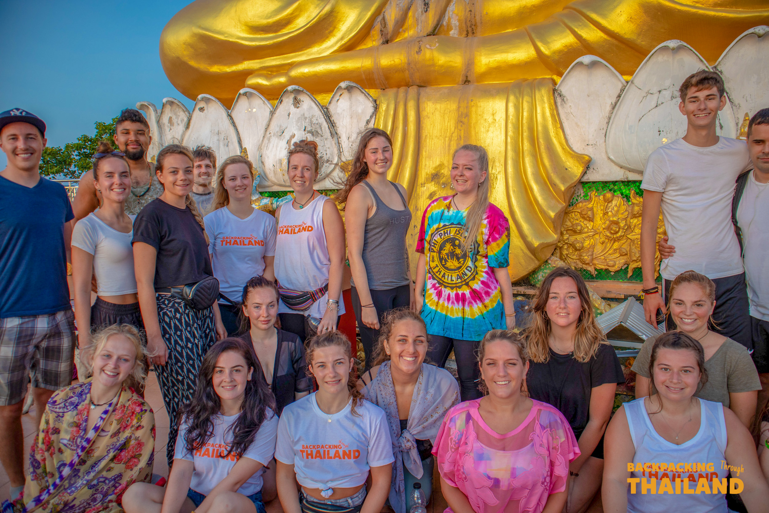 A large group of happy backpackers pose in front of a giant golden Buddha statue at Tiger Cave Temple.