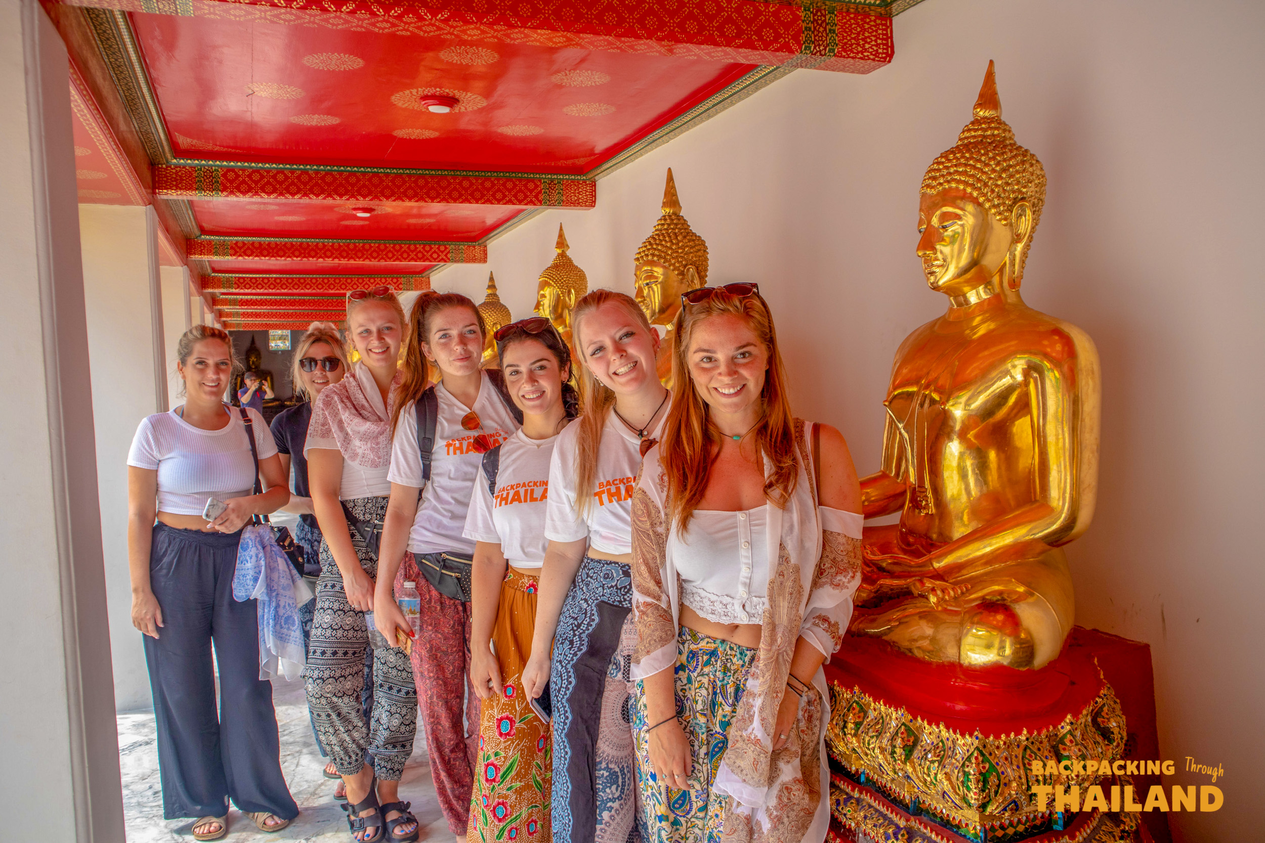 Seven happy backpackers pose with golden Buddha statues inside a beautiful temple in Bangkok.