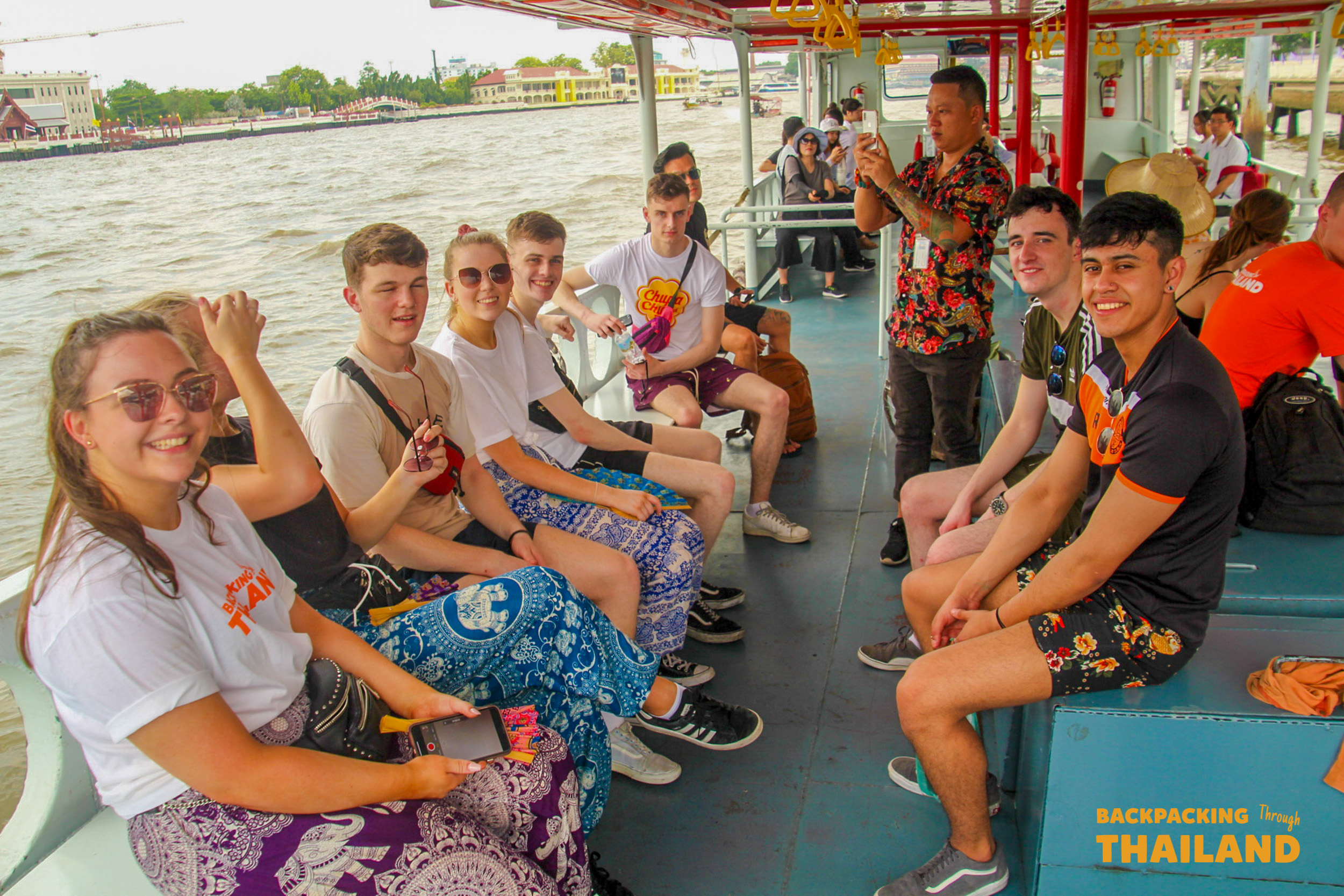 Smiling backpackers on a river boat tour in Bangkok, Thailand, listening to their guide.