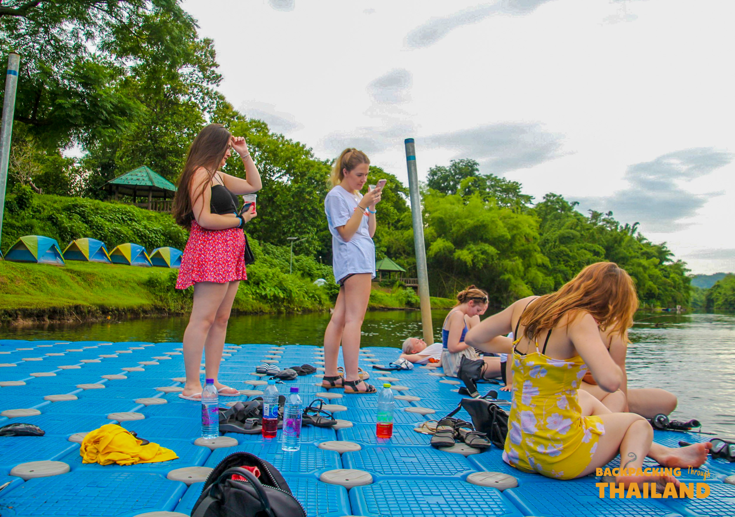 Backpackers relax on a floating platform by the Khwae Yai River with colorful tents on the bank.
