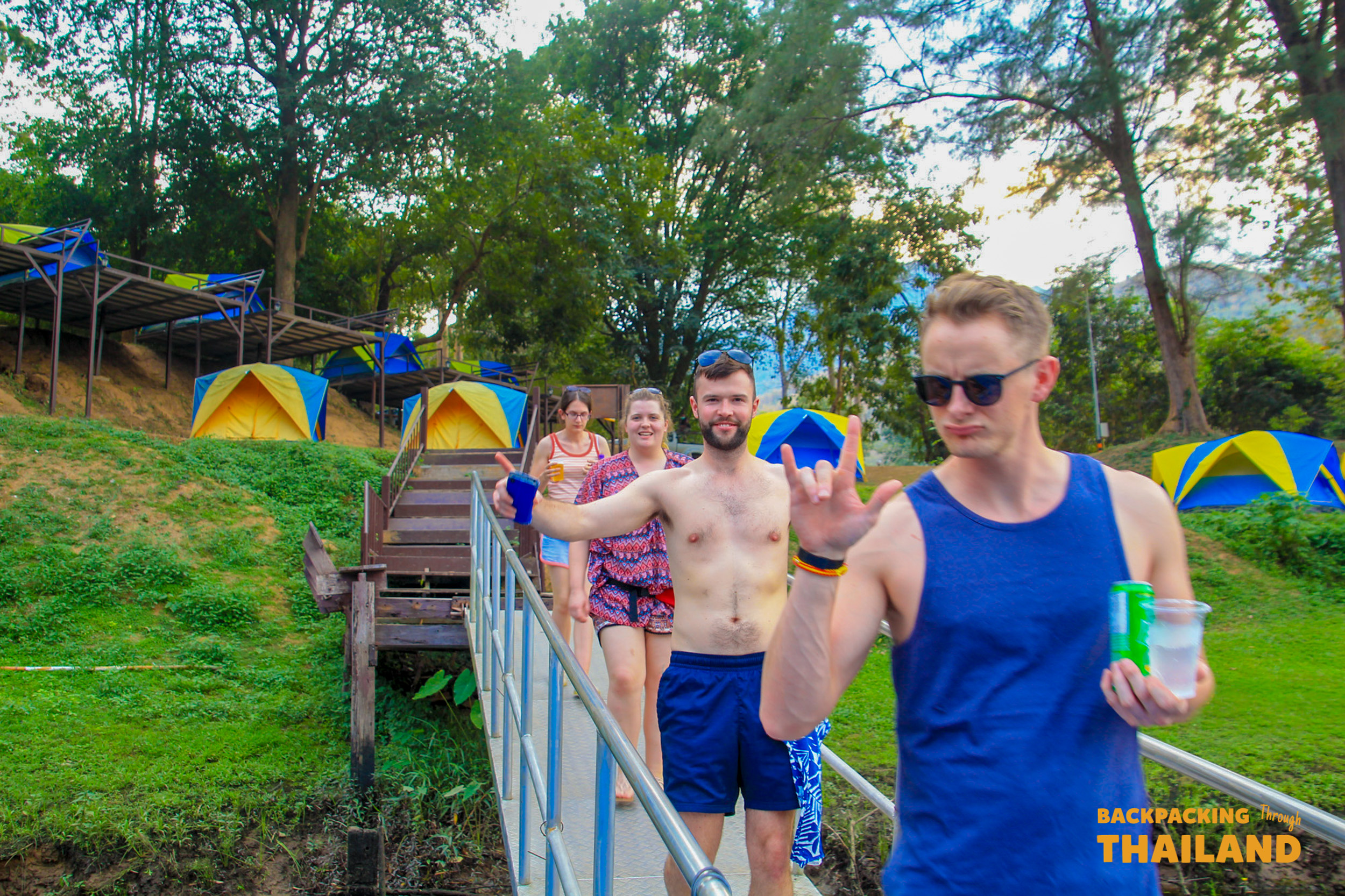 Smiling backpackers walk past their colourful tents at a relaxing campsite in Erawan National Park, Thailand.