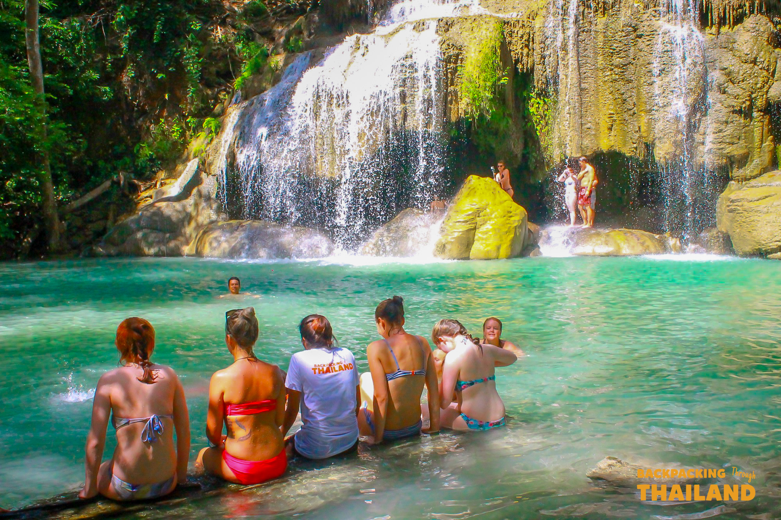 Backpackers relax in the emerald pools of Erawan Waterfalls in Thailand, enjoying the refreshing cascade.