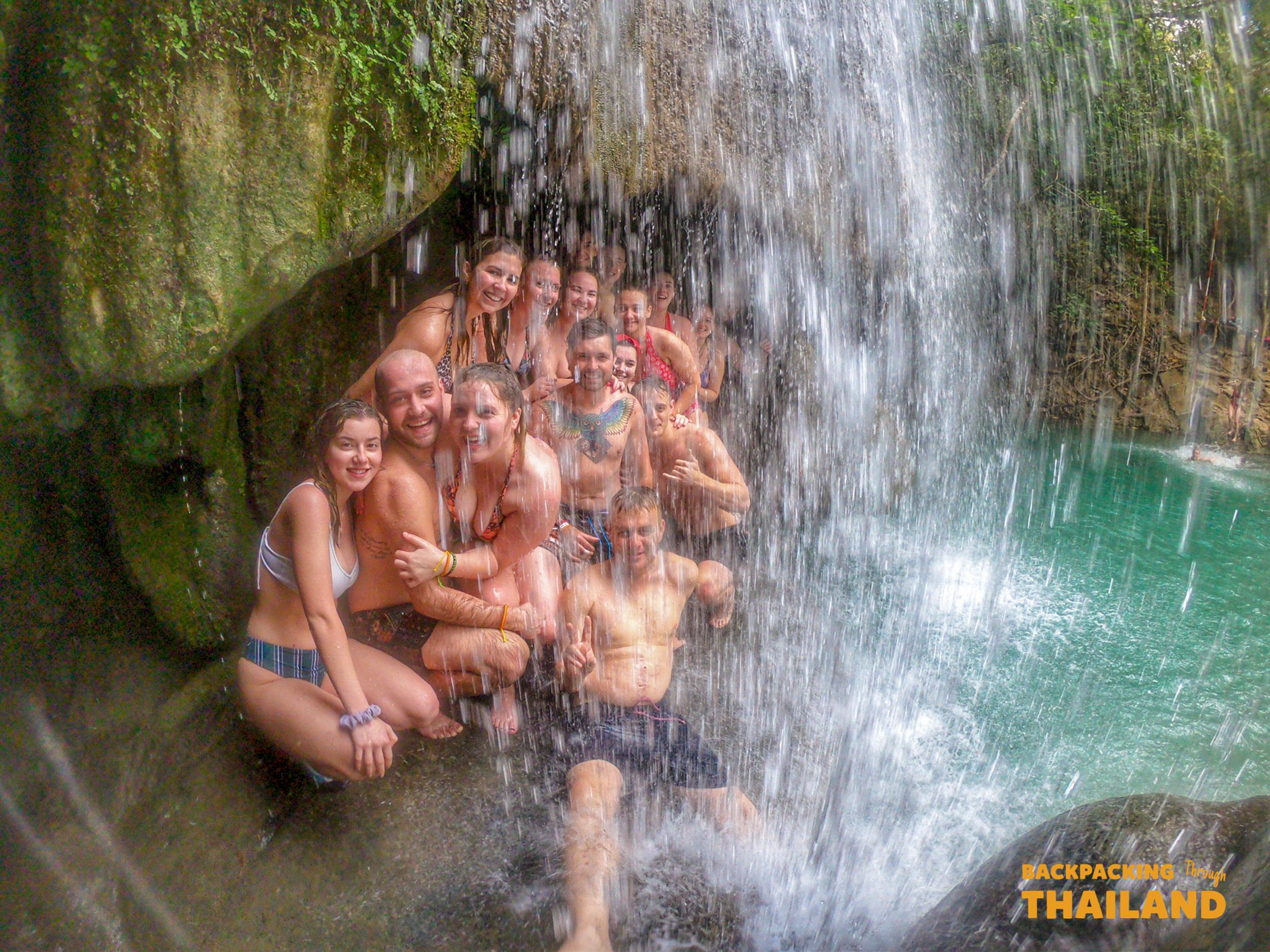 Smiling travelers cool off under the cascading waters of Erawan National Park in Thailand.