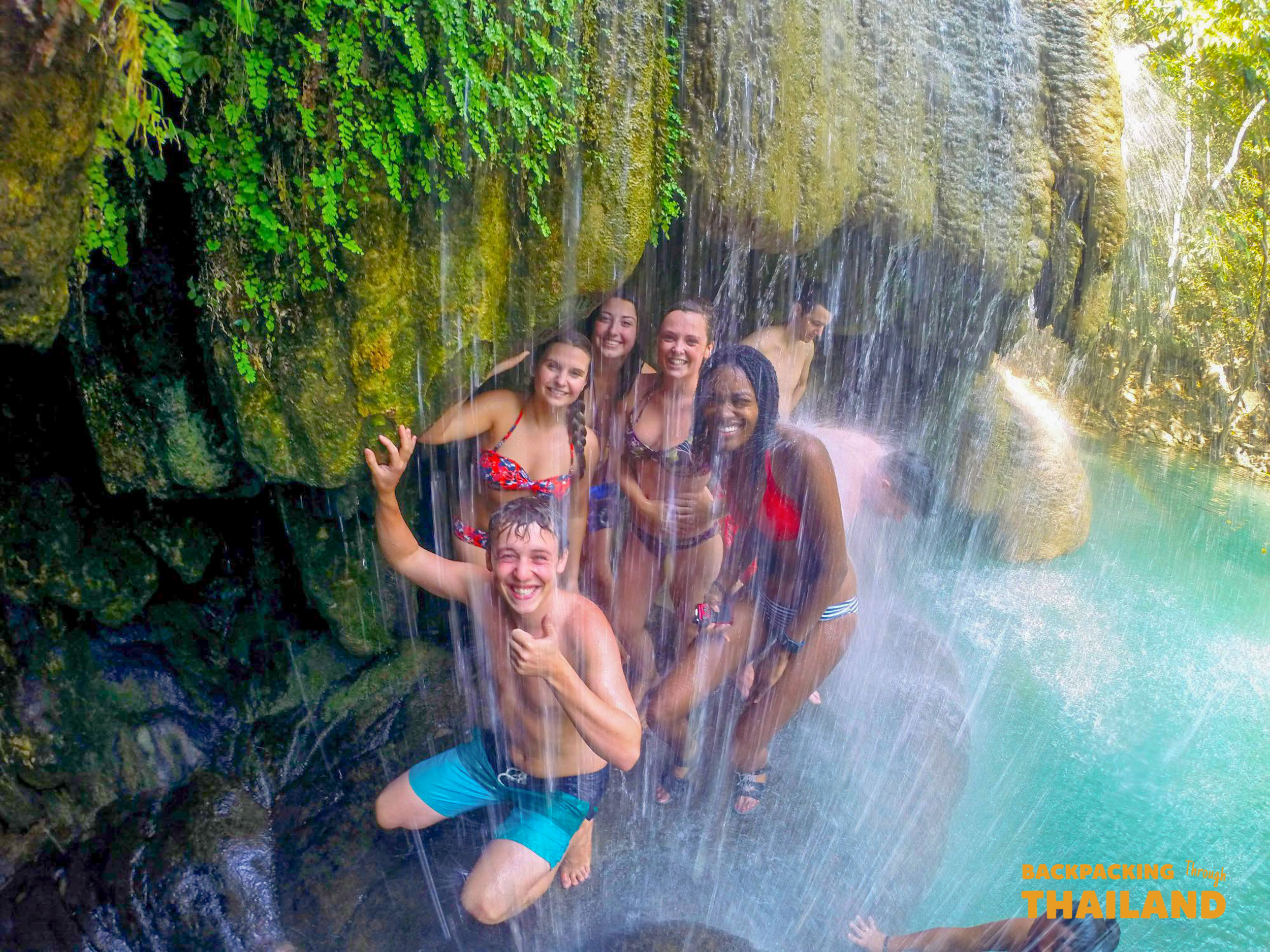 Six smiling friends cool off under the cascading waters of Erawan National Park, Thailand.