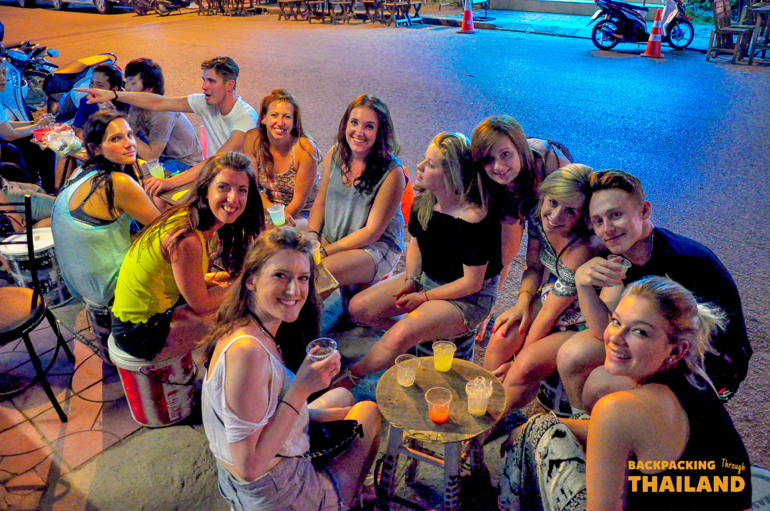 A group of happy backpackers enjoying drinks and conversation at a lively outdoor bar at night.
