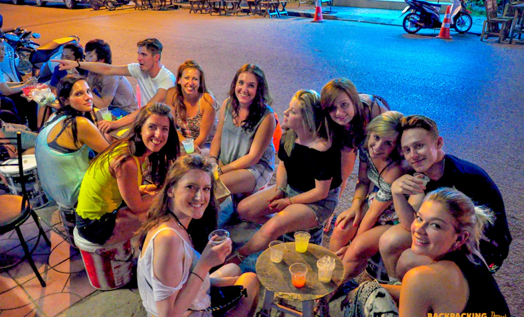 A group of happy backpackers enjoying drinks and conversation at a lively outdoor bar at night.
