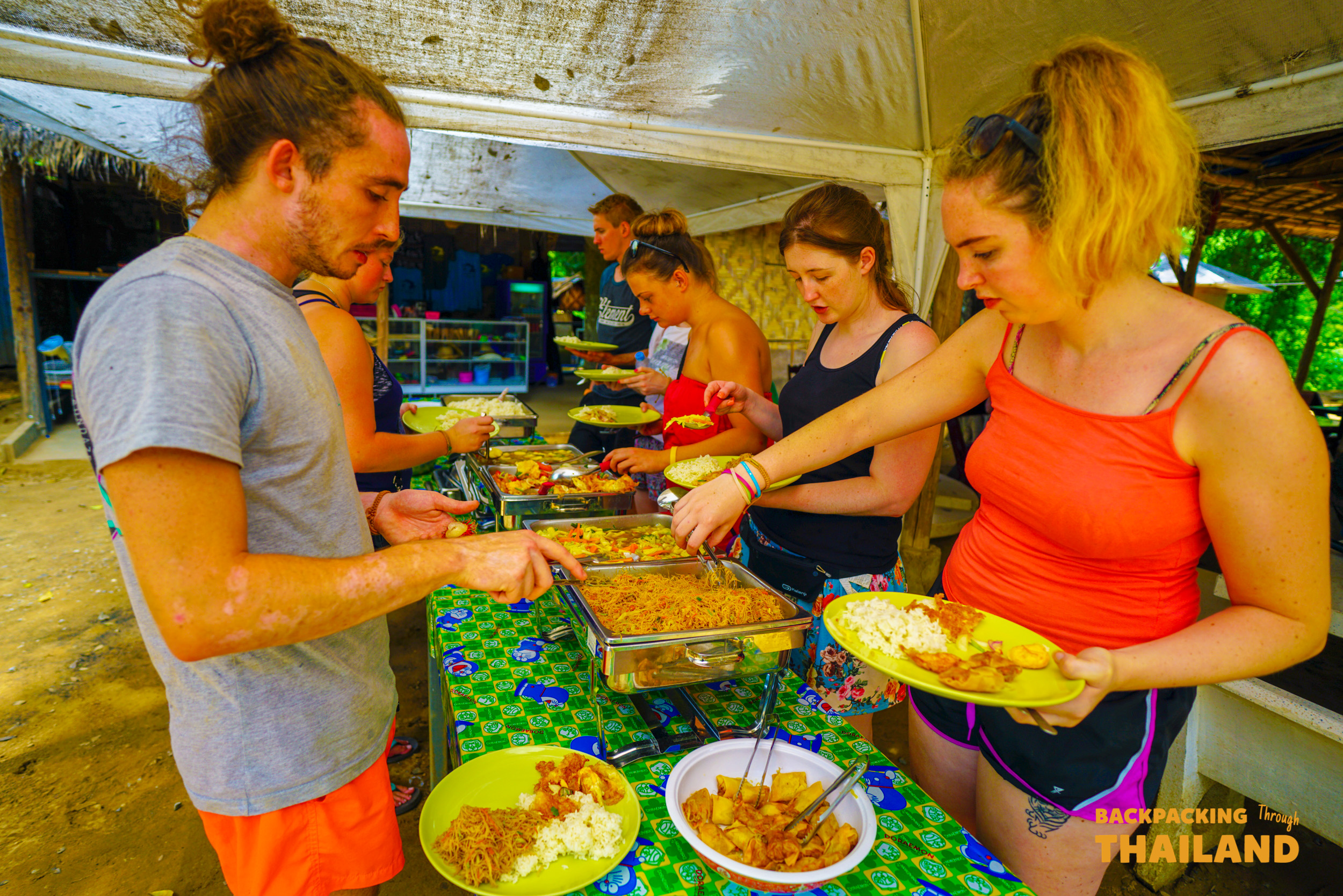Travelers fill their plates with various dishes from a buffet line in a relaxed outdoor setting.