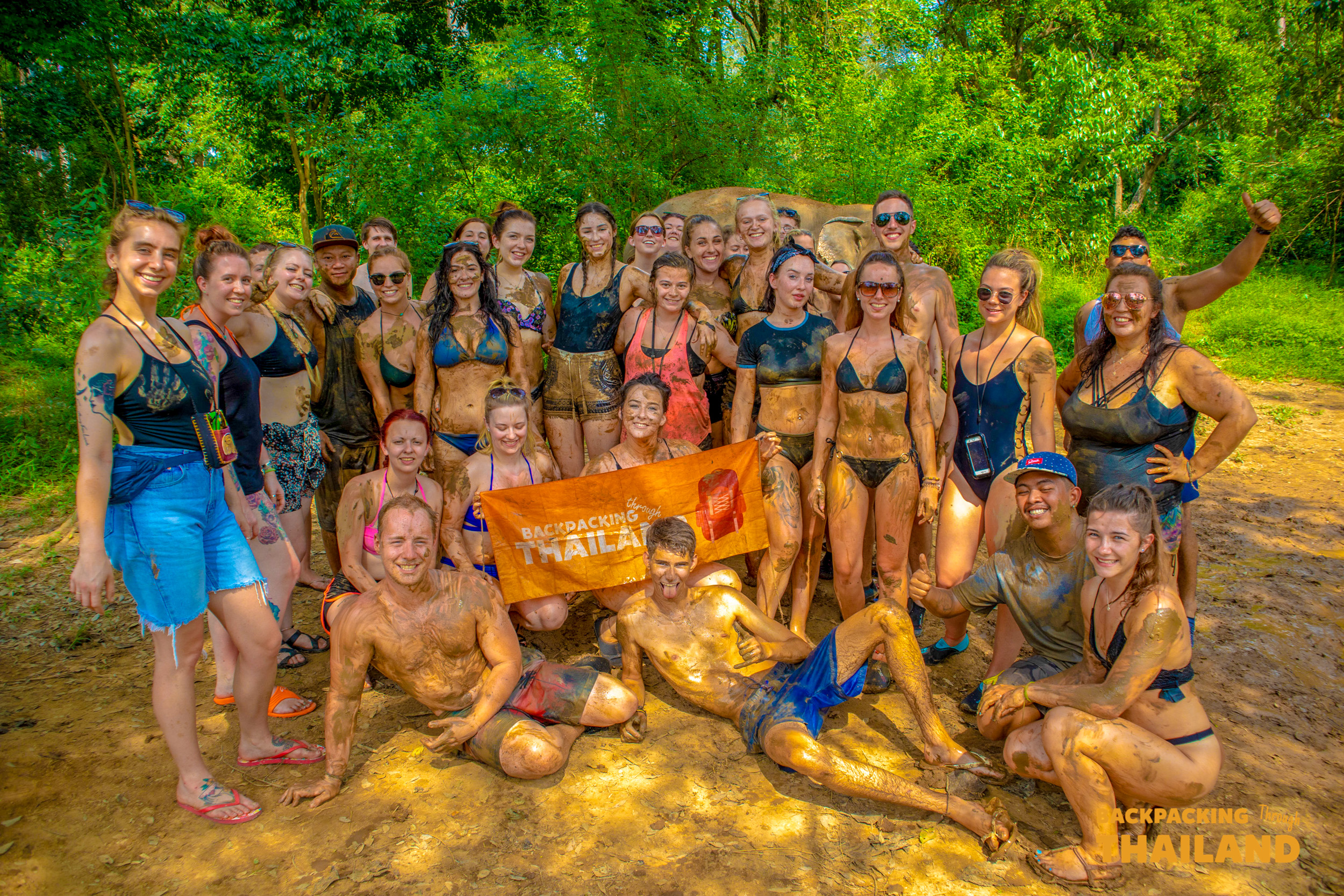 Happy backpackers covered in mud pose with an elephant at a sanctuary in Thailand.