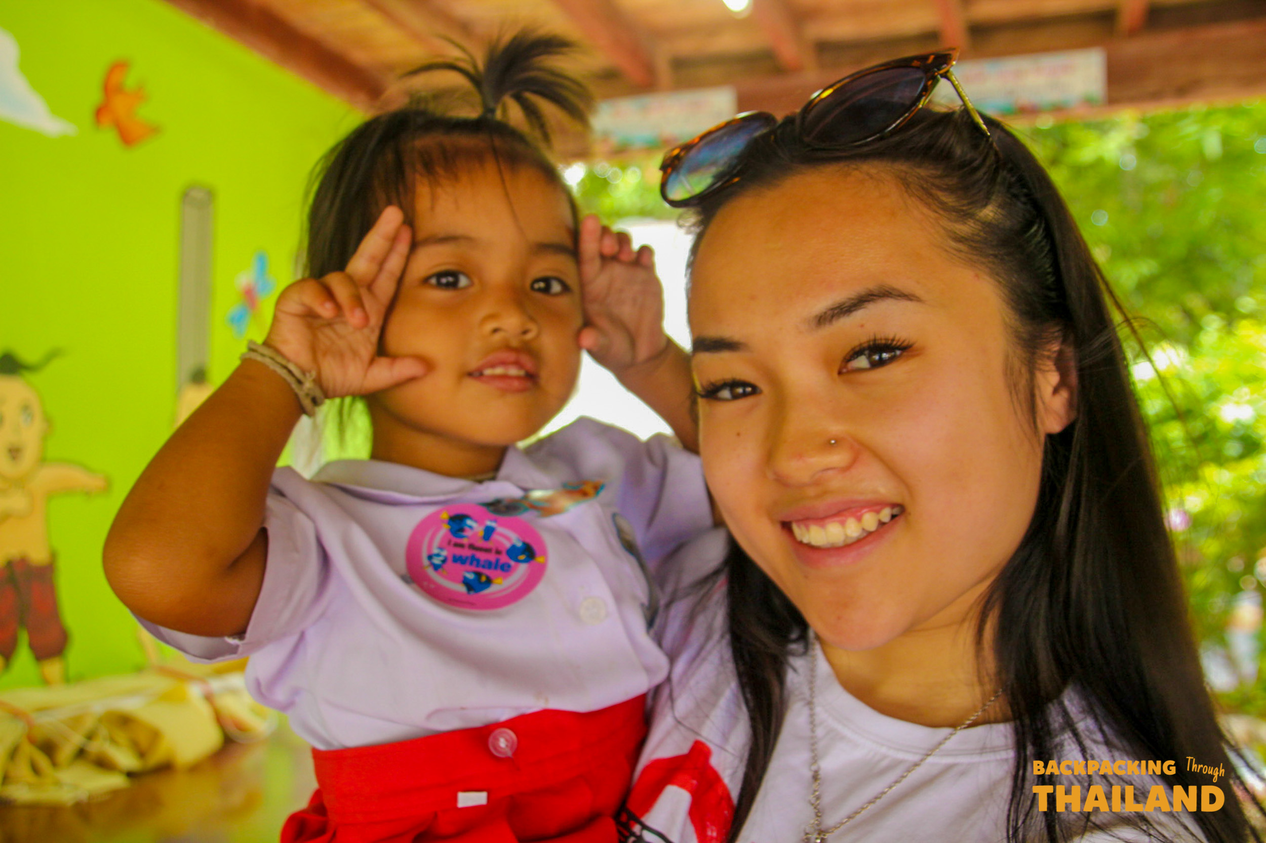 A smiling backpacker poses with a cheerful Thai schoolgirl making a peace sign at a local school.