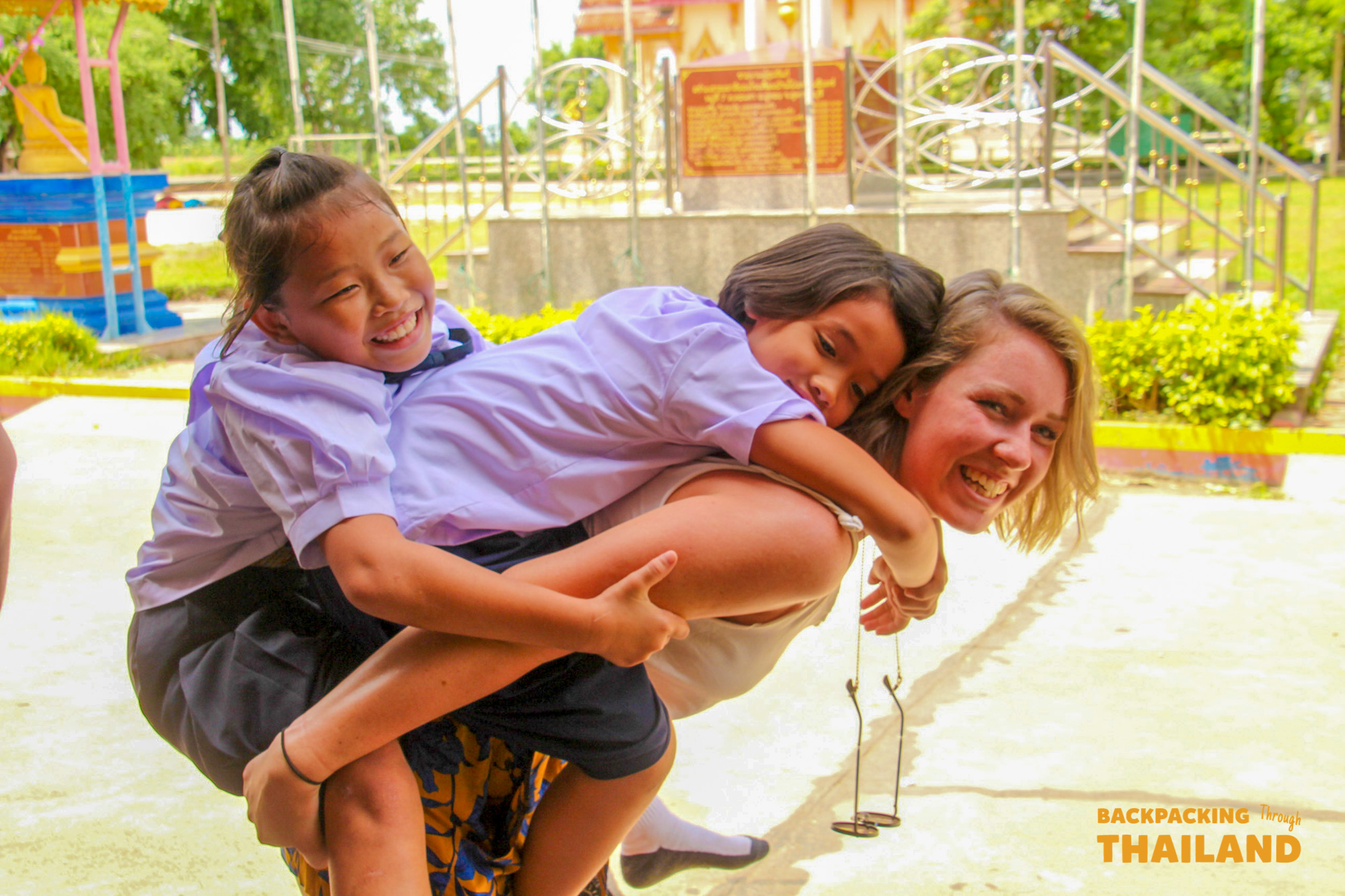 A joyful backpacker gives two smiling Thai schoolgirls a piggyback ride during a local school visit.